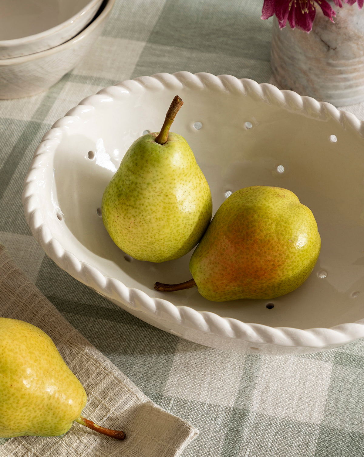 Two ripe pears sit in the Solene Stoneware Bowl with a braided rim on a checkered cloth. Another pear nearby, plus a glimpse of a flower vase, add charm to your kitchen decor.