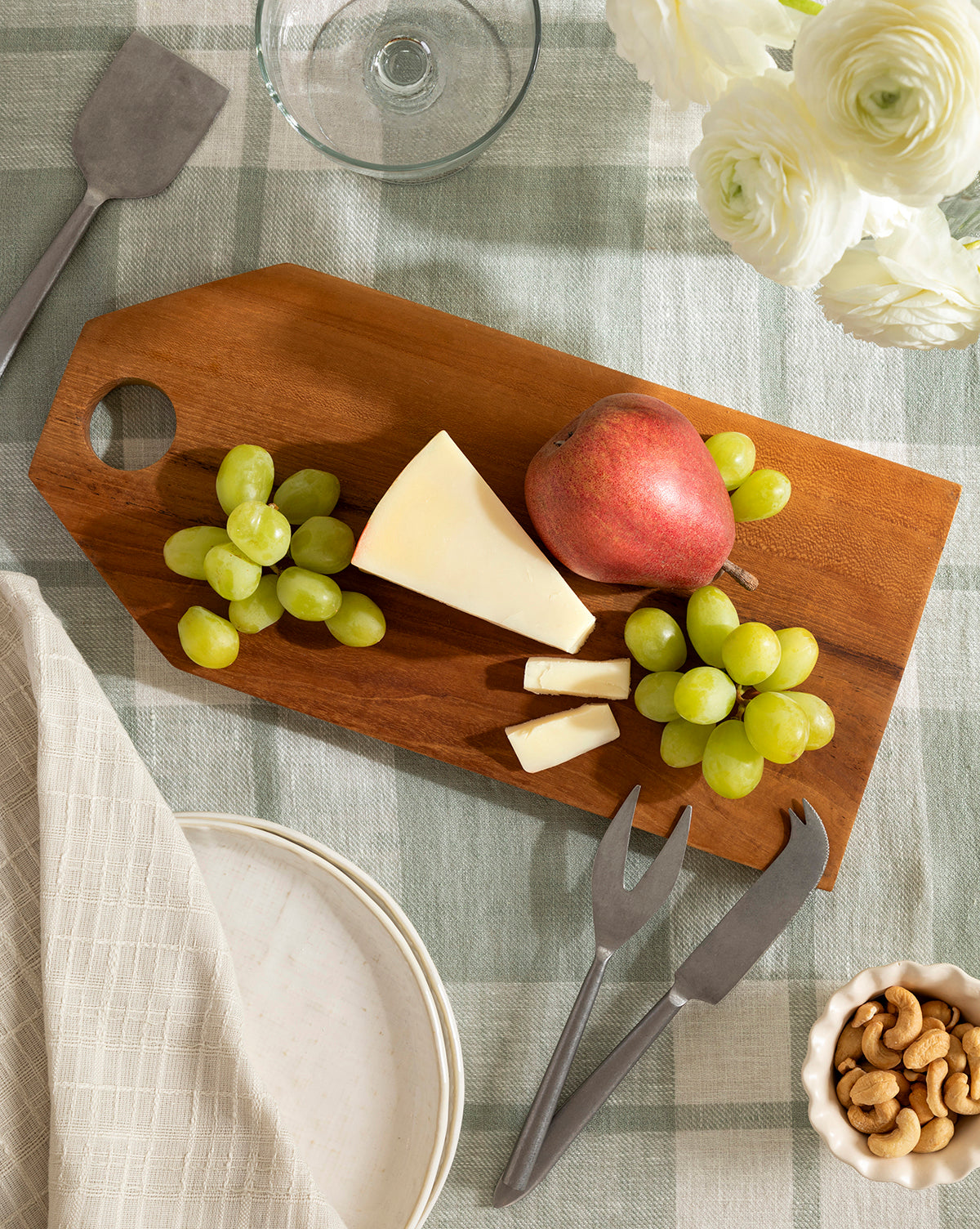 The Bevelled Edge Cutting Board holds green grapes, cheese with cheese forks, and a red pear on a checkered tablecloth. Nearby are cashews, white flowers, plates, and a napkin—making it ideal for any kitchen staple by McGee & Co.