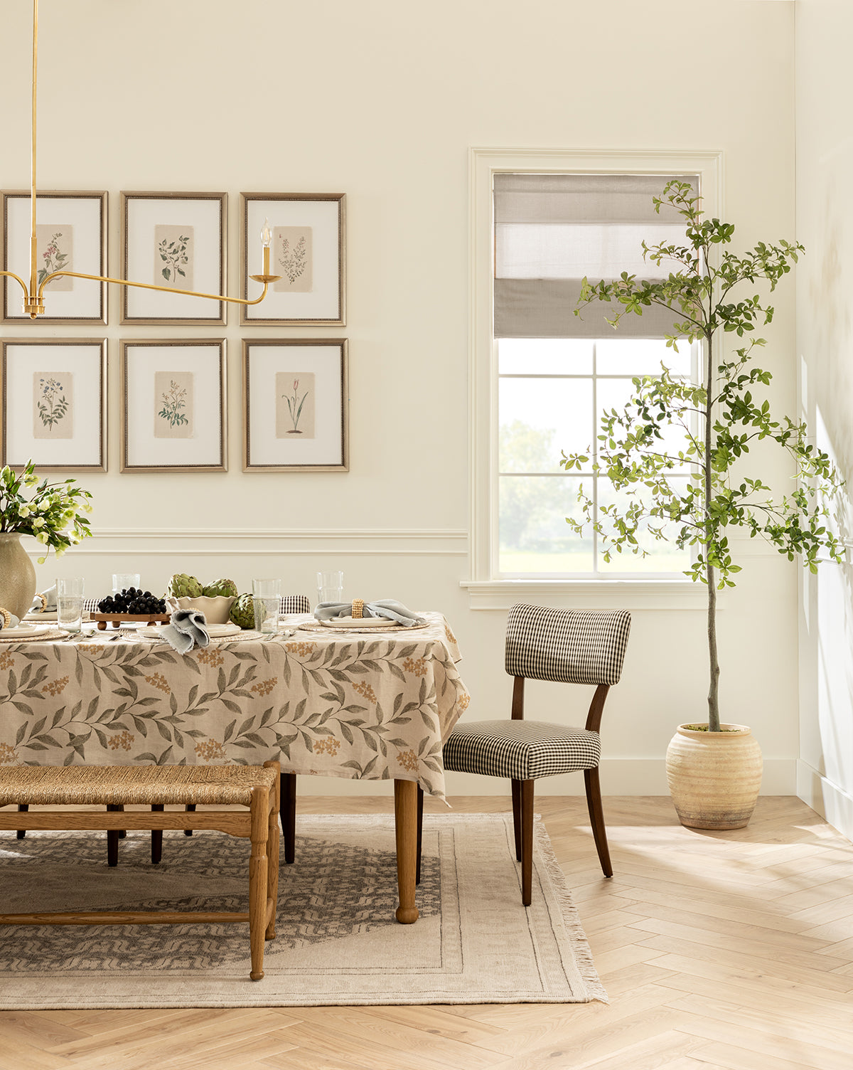 A bright dining room features a wooden table with a Petite Floral I tablecloth, chairs, a potted tree by the window, framed botanical giclee prints on the wall, and a gold chandelier overhead, by McGee & Co.