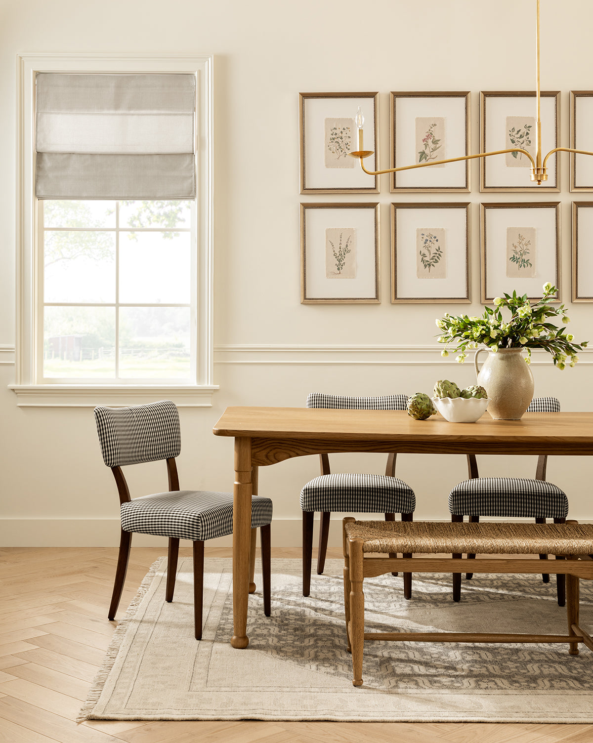 A bright dining room features a wooden table, plaid chairs, a woven bench, greenery in a vase, a light rug, the Petite Floral III wall art in a vintage wood frame, a brass light fixture, and a window with a gray Roman shade by McGee & Co.