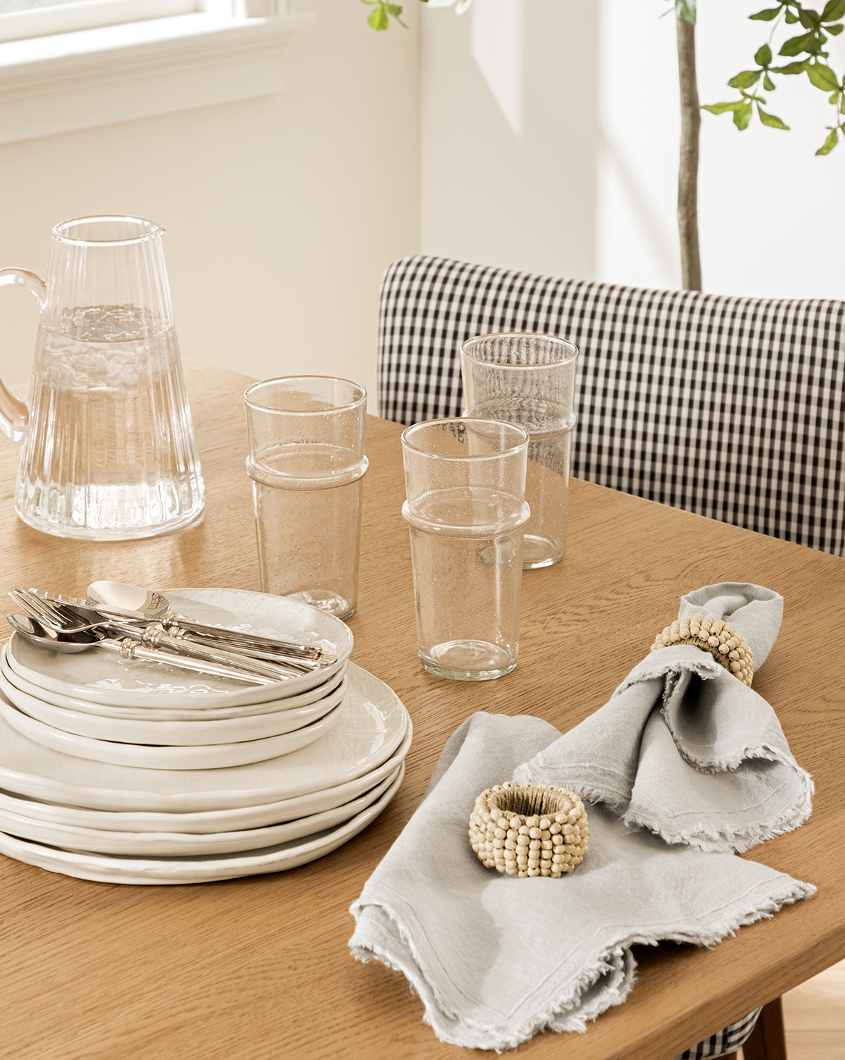 A wooden dining table set with white plates, silver cutlery, clear glasses, the Adonis Ribbed Glass Pitcher filled with water, and two gray napkins with beaded rings. Sunlight illuminates elegant kitchen styling and a checkered chair in the background, by McGee & Co.