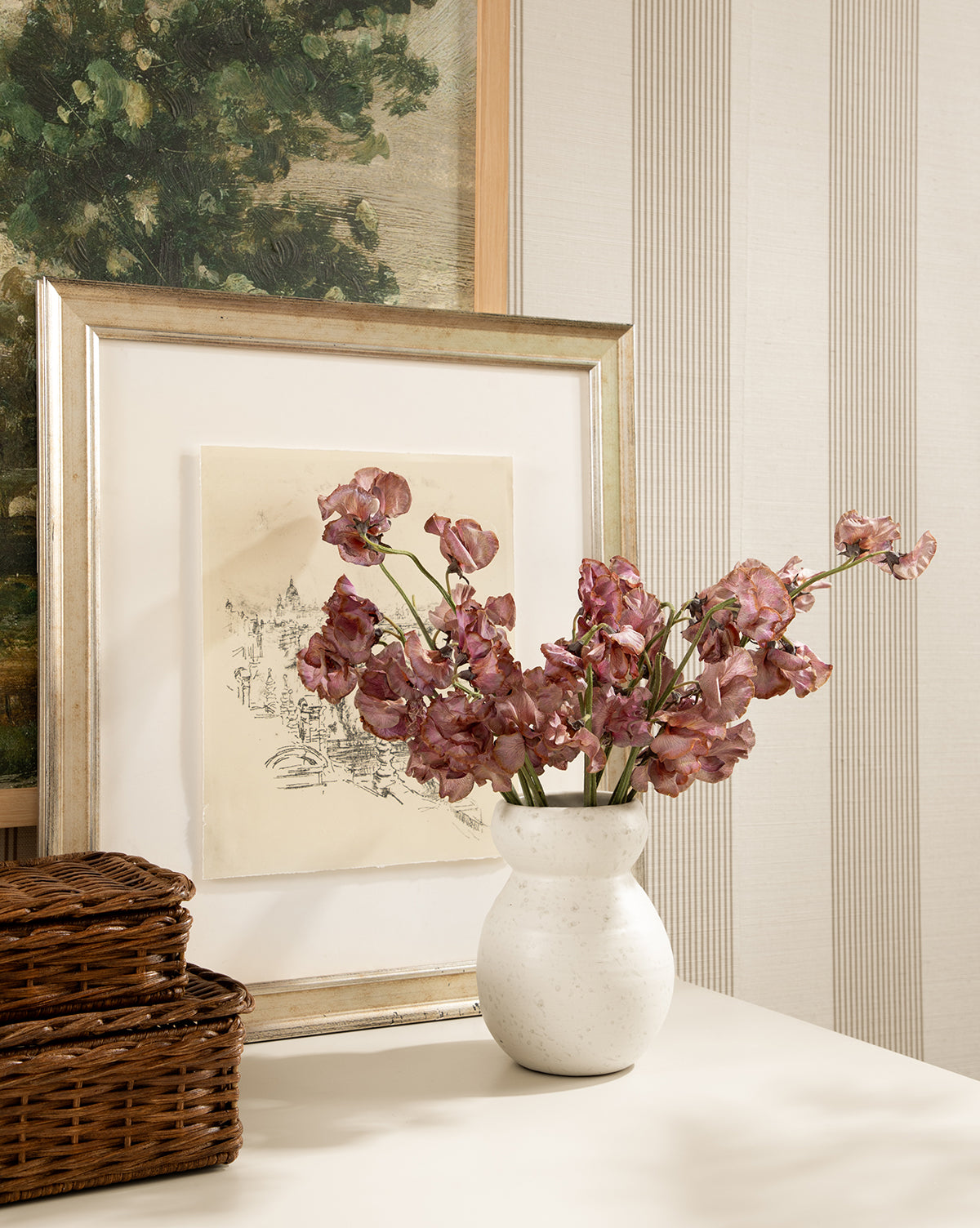 The Lyra Vase, white ceramic with mauve flowers, sits on a table beside two woven baskets. Behind it are framed pictures—one of a tree, the other a building sketch—all set against striped wallpaper, by McGee & Co.