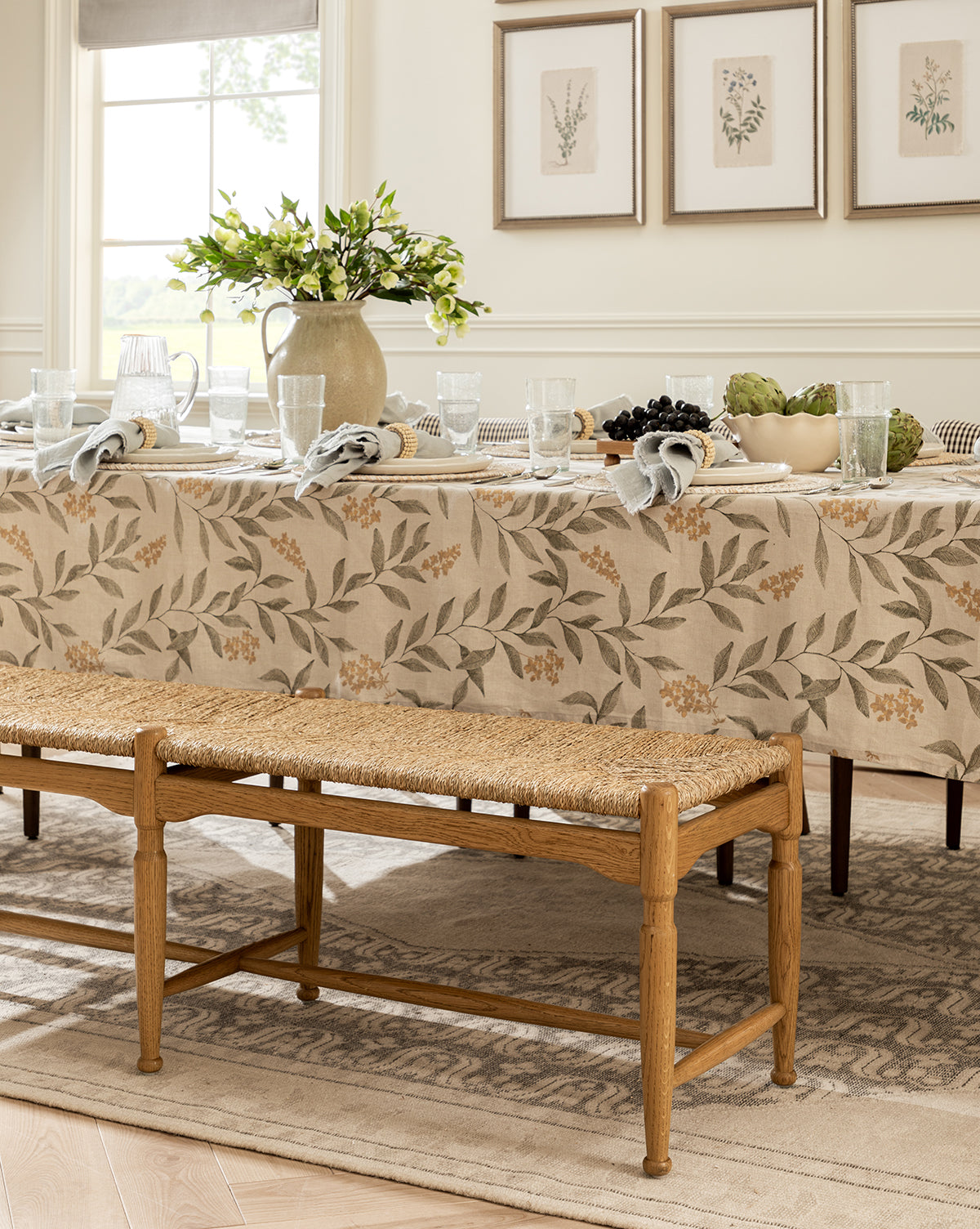 A dining table with a floral tablecloth is set with plates, glasses, napkins, and a ceramic jug holding green branches. The room features the Phipps Woven Bench and framed botanical prints for an elegant, neutral-toned look by McGee & Co.
