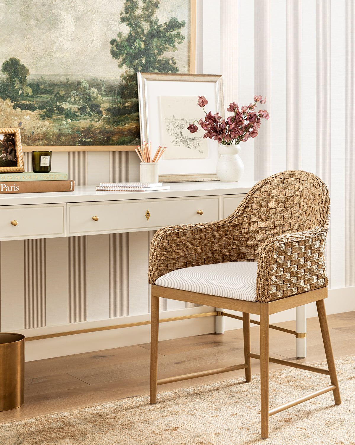 The Molly Arm Chair with a white cushion is placed before a white desk with gold drawer pulls. On the desk are pink flowers in a vase, framed art, books, and pencils. A landscape painting hangs above the setup by McGee & Co.