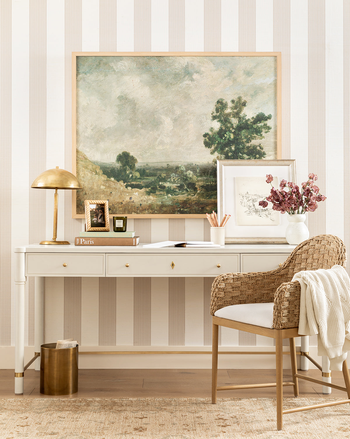 A stylish workspace with a white desk, gold lamp, books, pink flowers in a vase, and a wicker chair with cushion. The striped wallpaper highlights the English Meadows giclee on canvas wall art in a natural oak frame, by McGee & Co.
