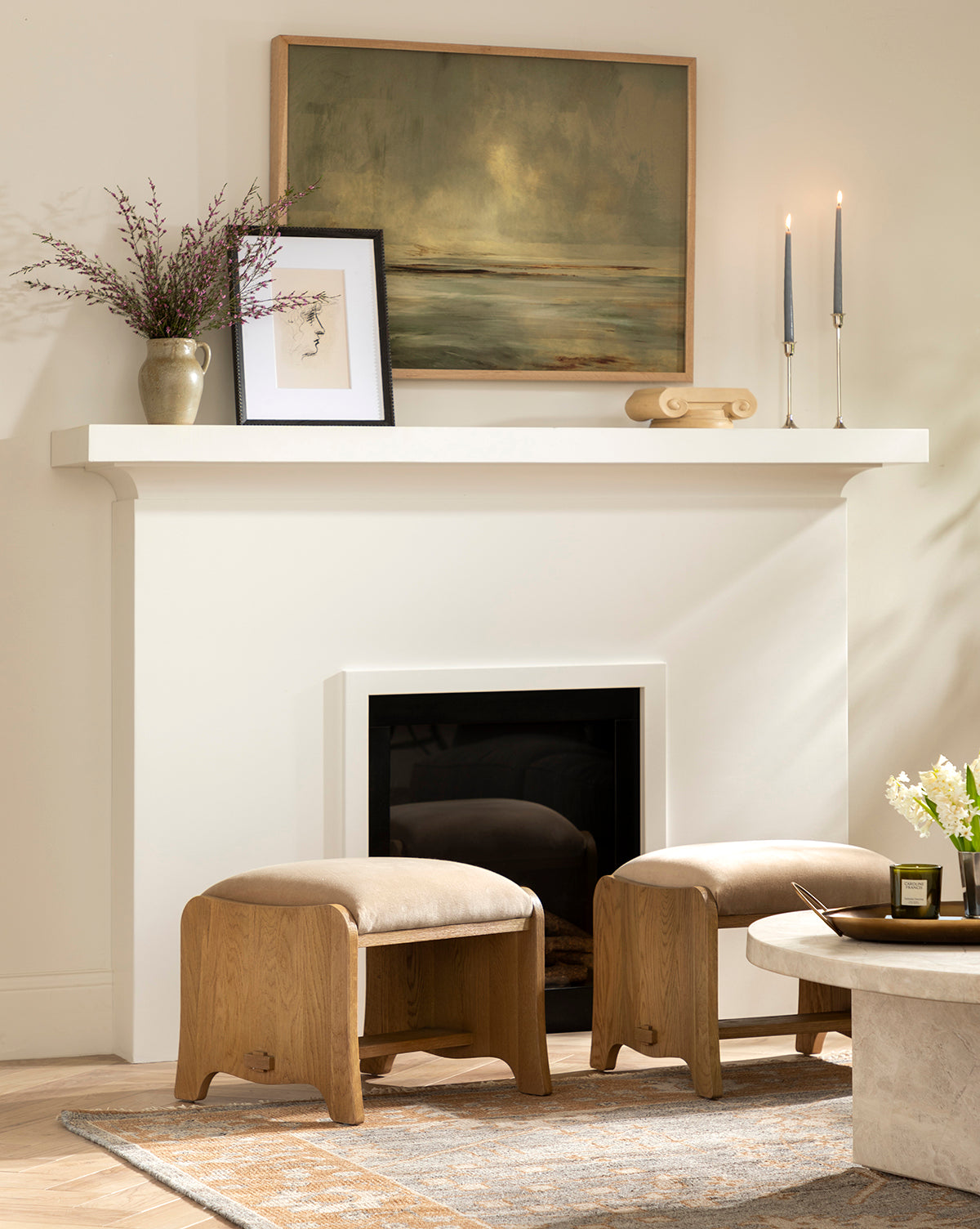 A modern living room with a white fireplace mantle, featuring the Misty Blue Headland giclee print in a white oak frame, a vase with branches, tall candles, wood decor pieces, two wood stools nearby, a round table, and a rug by McGee & Co.