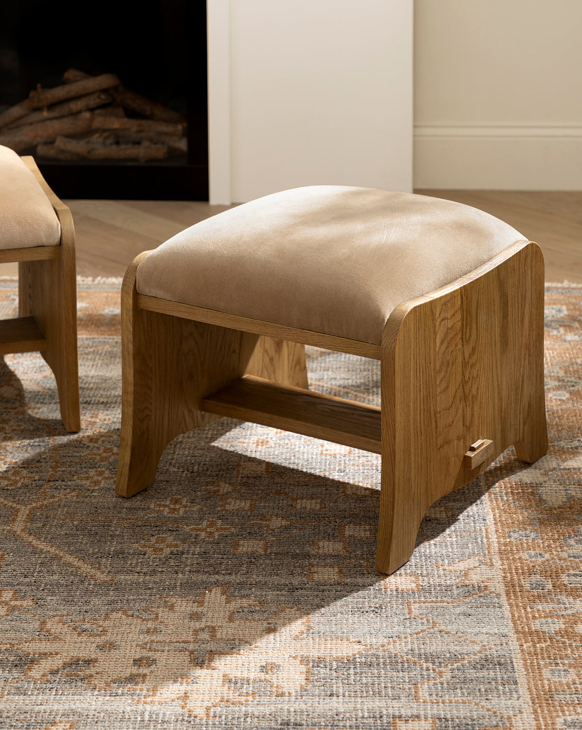 A Makerspalm Gable Ottoman in Drift Oak with a Fawn Velvet cushioned top rests on a patterned rug in a bright room featuring light walls and wood floors, McGee & Co.