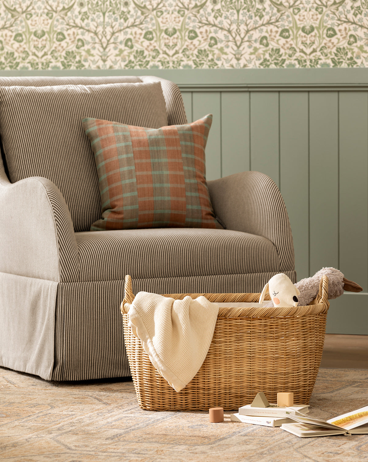 A striped armchair with a plaid pillow sits beside the Fairway Laundry Basket, filled with a blanket and stuffed toys. Wooden blocks and an open book are on the floor, while green paneling and floral wallpaper decorate the room by McGee & Co.