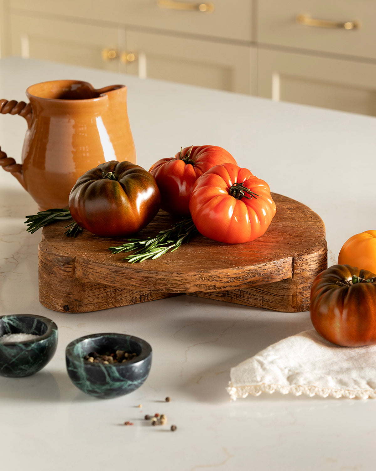 Heirloom tomatoes and rosemary rest on the Norfolk Wood Pedestal, with a brown ceramic pitcher, two marble bowls, and a folded cloth enhancing the home decor in the background, by McGee & Co.