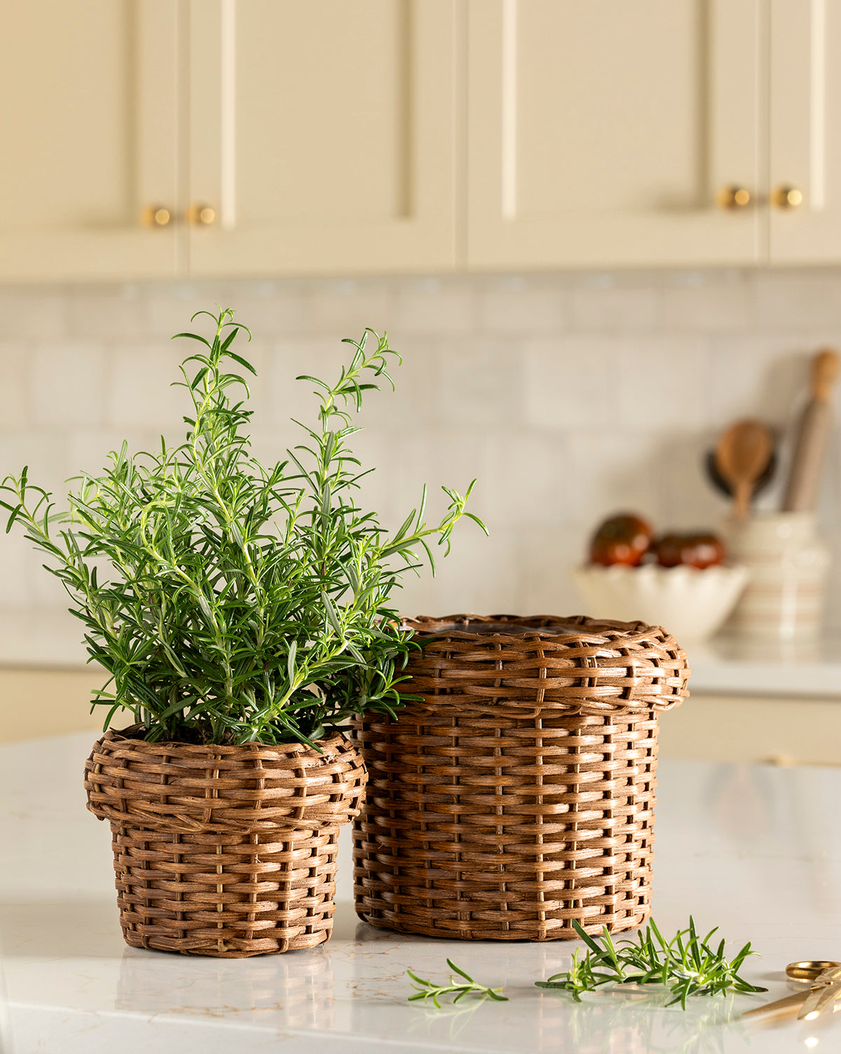 The Huger Woven Planter by DURUS INDUSTRIES CORPORATION, featuring a rosemary plant, sits next to a larger empty wicker basket on a white kitchen countertop with blurred cabinets and utensils in the background—McGee & Co.