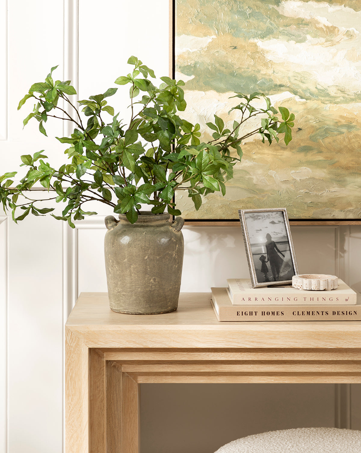 A green leafy plant in the Emilio Urn sits on a light wooden console table styled with vintage decor—stacked books, a small dish, and a framed black-and-white photo—beneath a painted landscape, by McGee & Co.