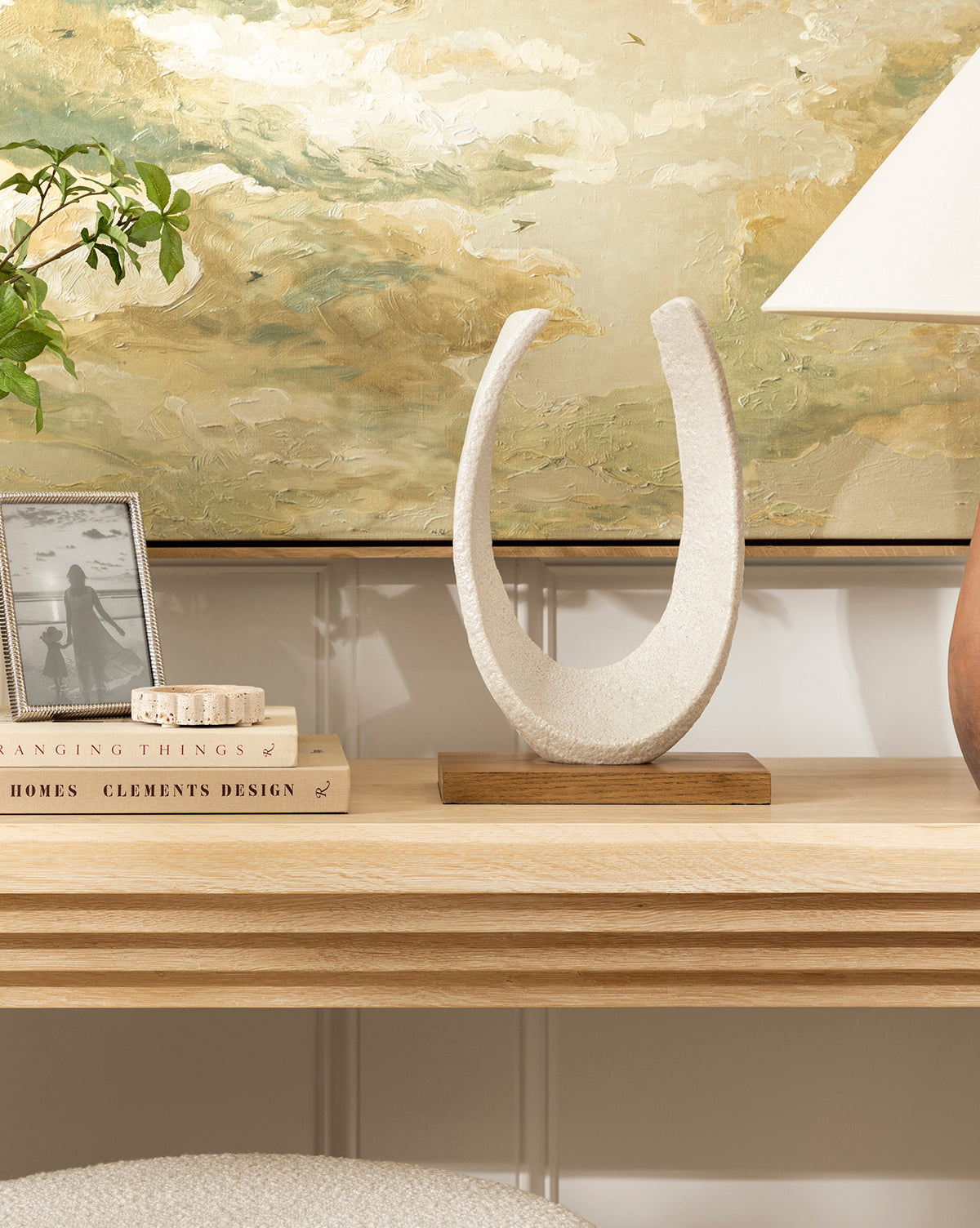 A light ash wood console table displays the sculptural Sergio Object on a wooden base, along with a lamp, framed photo, books, and plant, all set against an abstract earth-toned painting for chic contemporary style.