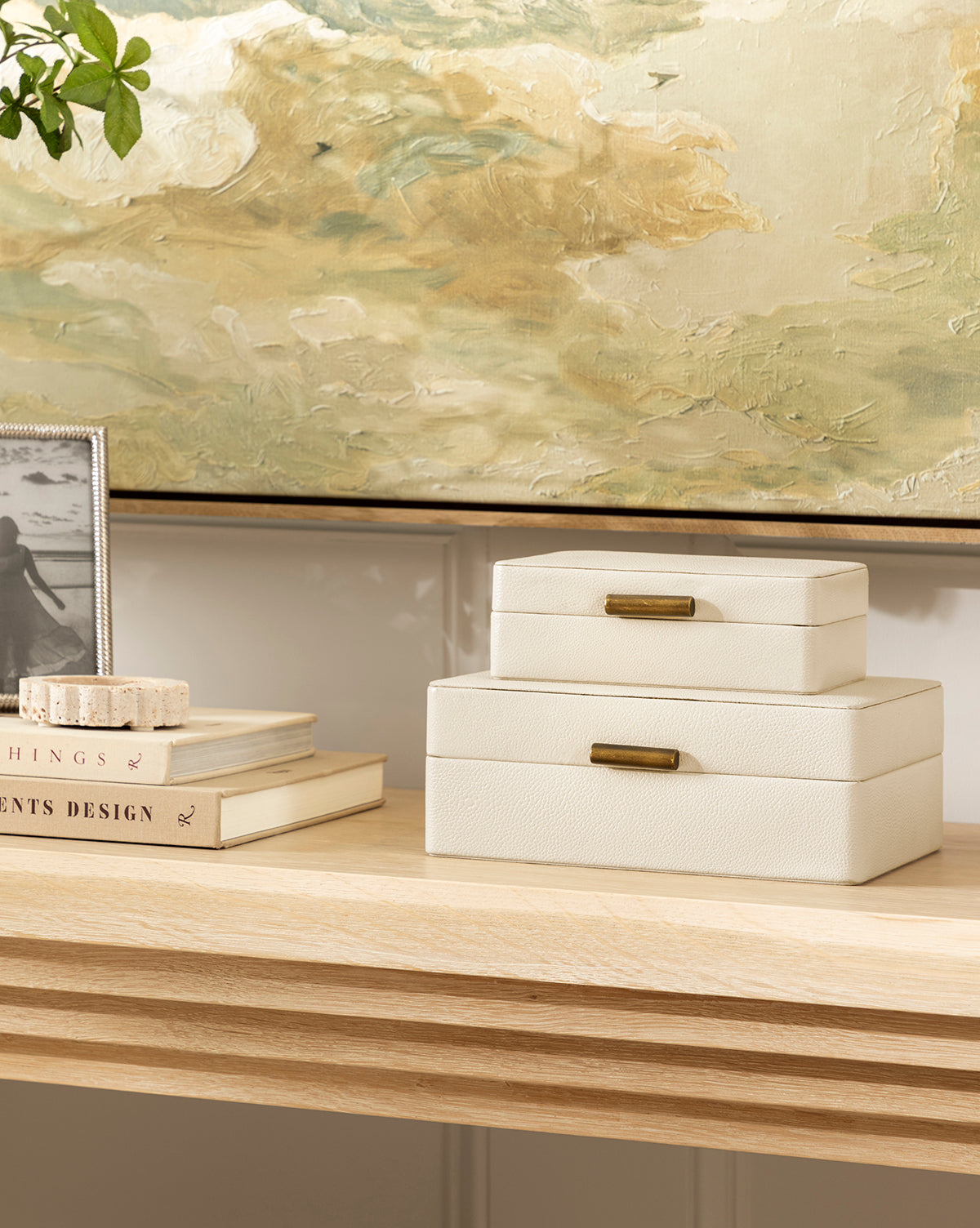 Two stacked Cream Shagreen Boxes with brass handles sit on a light wood table beside a photo frame, books, and a painted artwork featuring soft green, cream, and yellow tones in the background.