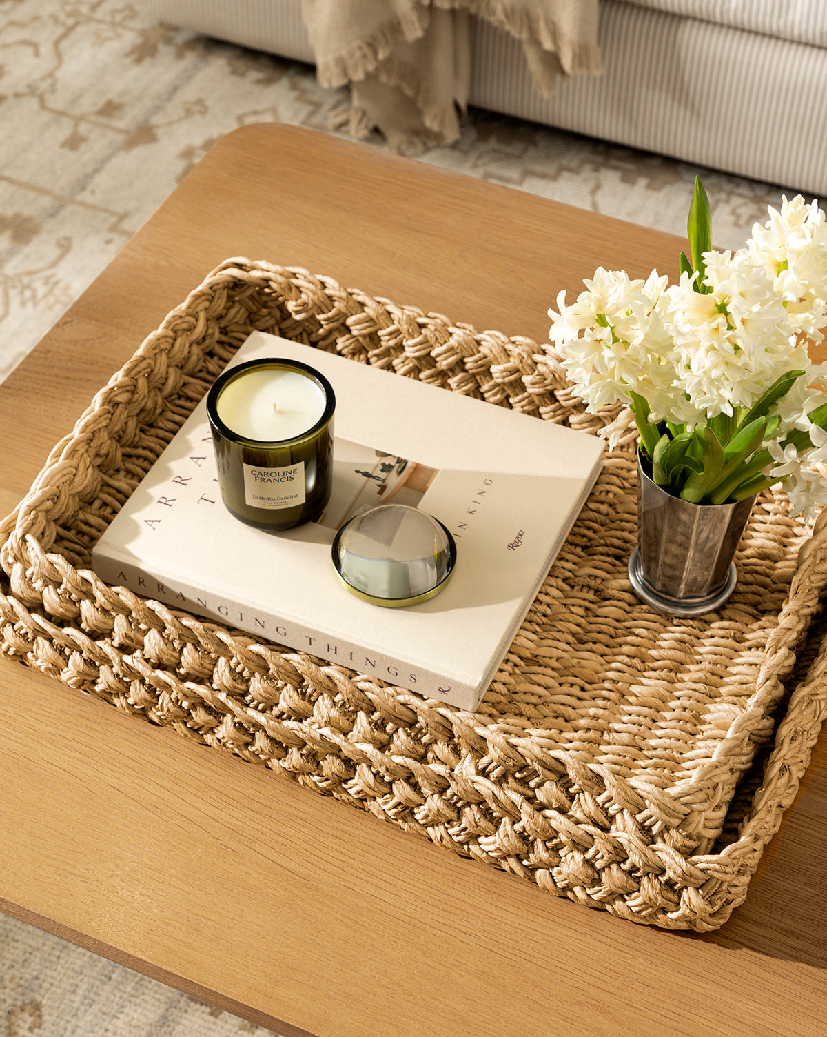 The Eudora Braided Trays (Set of 2) sits atop a wooden table, displaying a candle, closed book, and metal pot with white flowers; a cozy throw and sofa complete the inviting scene in the background, by McGee & Co.