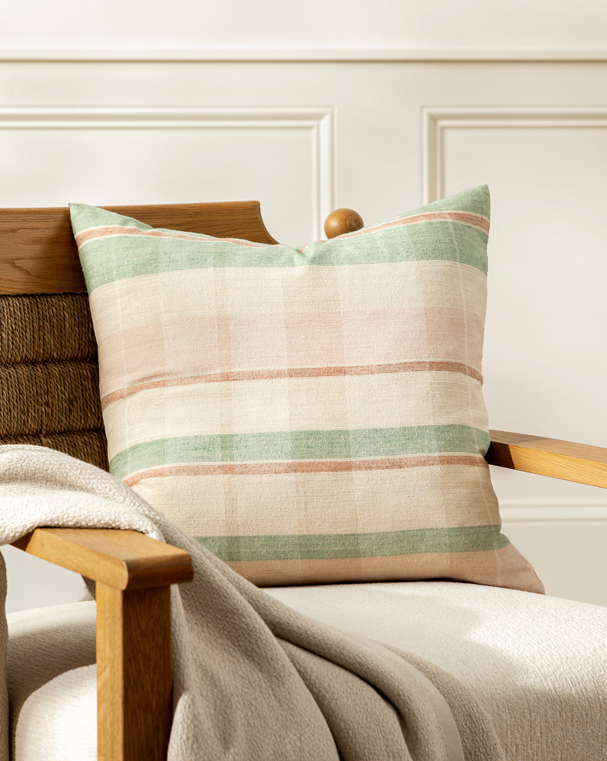 A wooden armchair with a beige textured blanket, topped with the Barret Pillow Cover—a hand-woven square pillow featuring pastel green, beige, and muted pink stripes—set against a white paneled wall, by McGee & Co.
