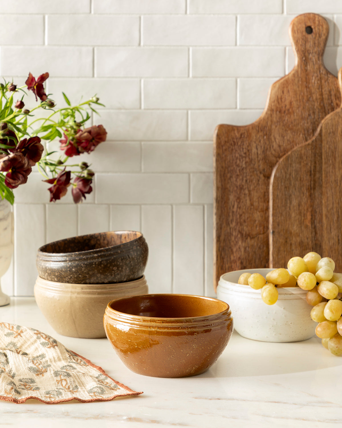 A set of four Mariella Stoneware Bowls in brown, beige, and white are displayed on a kitchen counter near grapes, wooden cutting boards, a patterned cloth, and a vase with dark red flowers against a white tile backsplash, by McGee & Co.