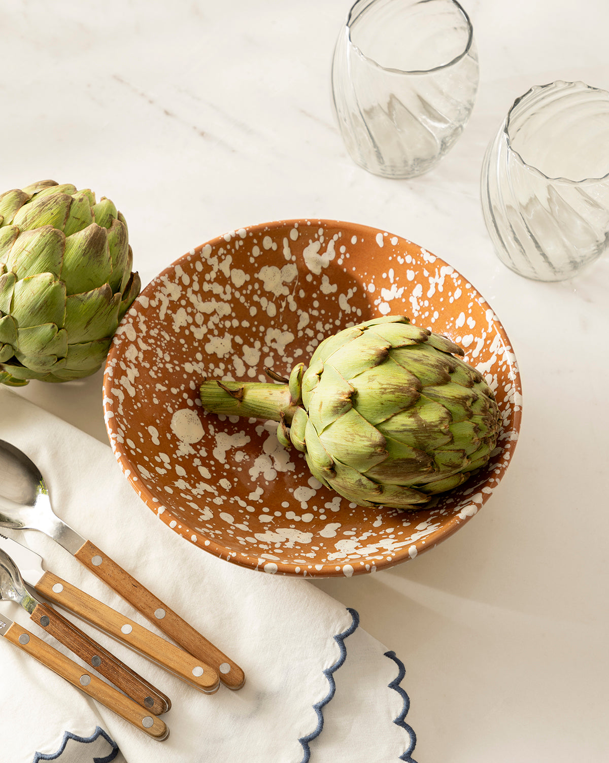 The Splatter Large Bowl, featuring a speckled brown ceramic design and holding an artichoke, sits beside another artichoke, three clear glasses, and wooden-handled utensils on a white napkin with blue trim on a white surface by McGee & Co.