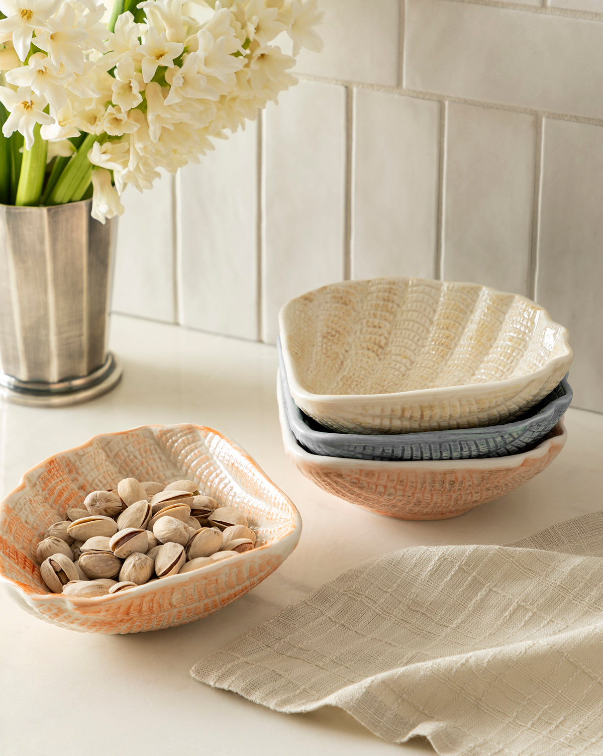 Four coastal-inspired Aurelie Shell Shaped Bowls are displayed on a countertop—one filled with pistachios, two stacked, and one beside them—with a beige cloth and metal vase of white flowers in the background against a tiled wall by McGee & Co.