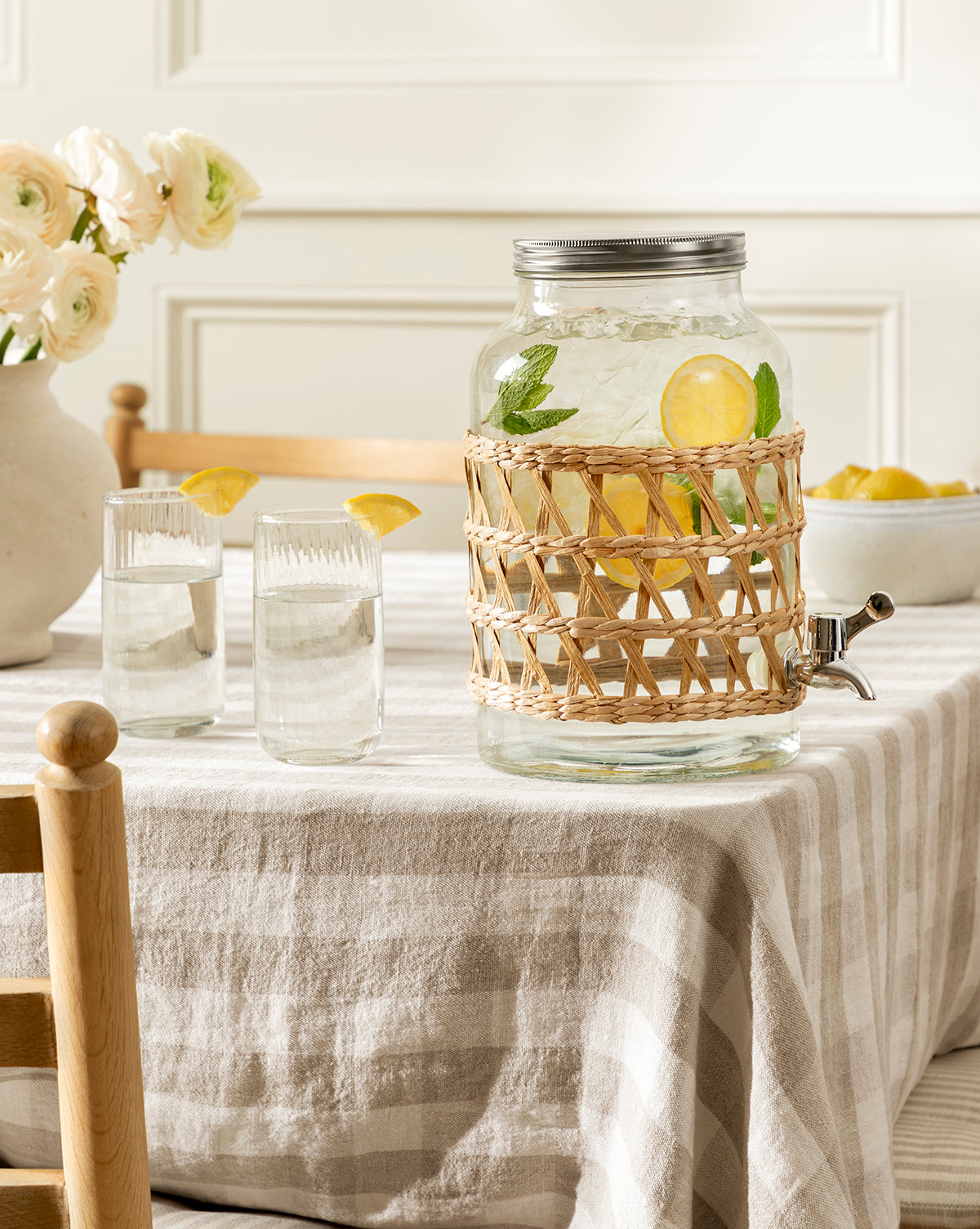 The Glass Beverage Dispenser with Woven Sleeve, filled with lemon slices and mint, sits on a striped tablecloth beside two glasses of water, a bowl of lemons, and a vase of flowers in the background, by McGee & Co.