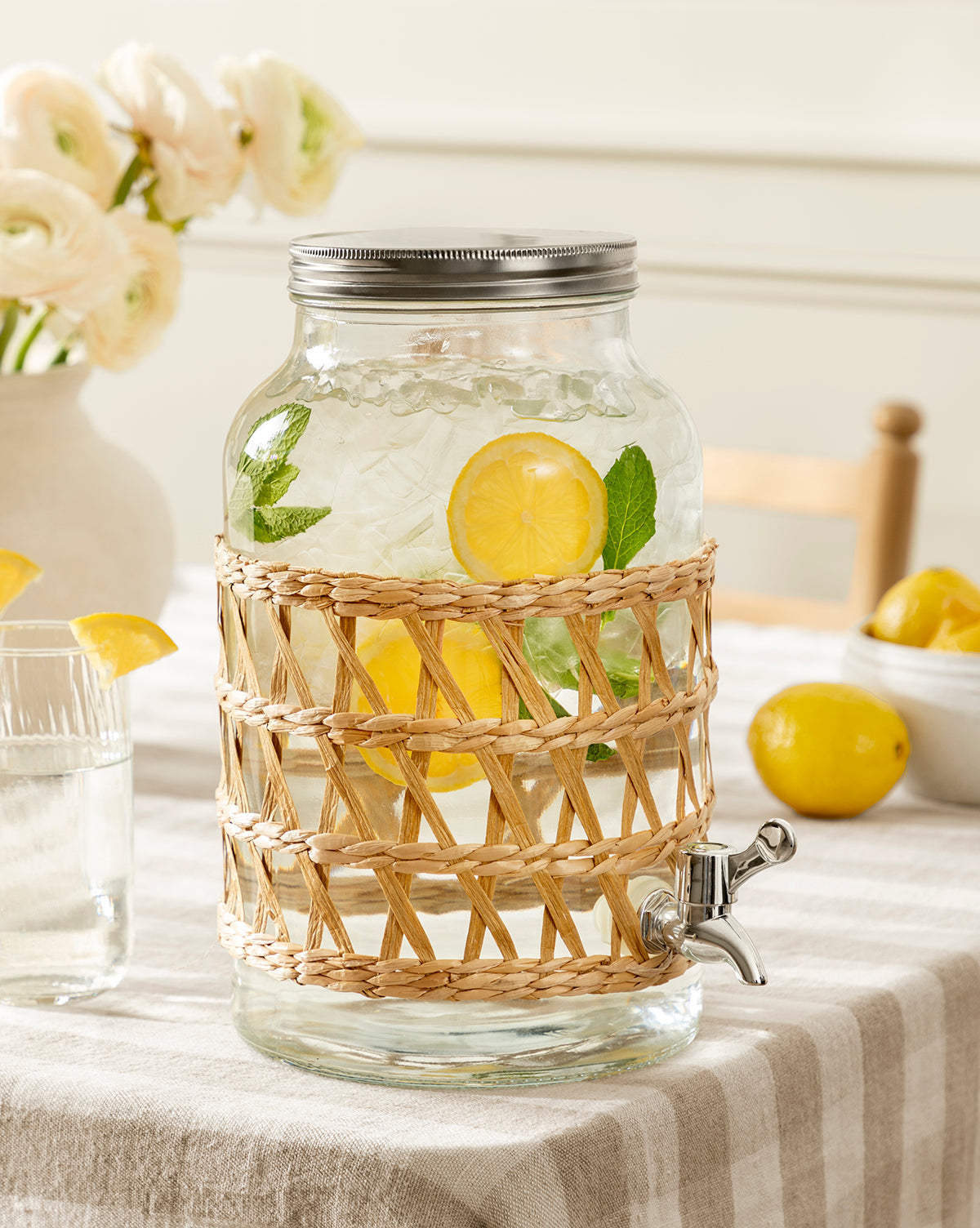 The Glass Beverage Dispenser with Woven Sleeve, filled with ice water, lemon slices, and mint, sits on a striped tablecloth alongside a glass of water and a bowl of lemons with flowers in the background, by McGee & Co.