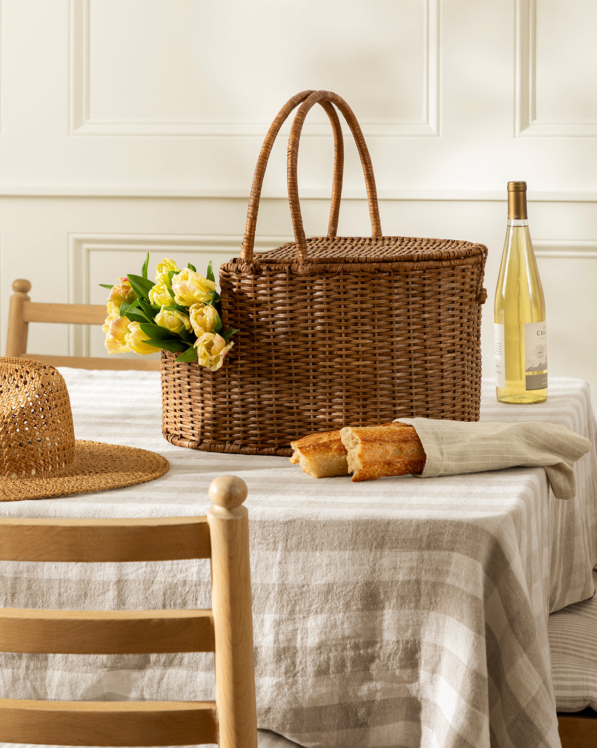 The Rattan Wicker Picnic Basket sits on a striped tablecloth next to a straw hat, baguette, and white wine. Surrounded by wooden chairs in a softly lit room, it offers an inviting and stylish storage solution for gatherings by McGee & Co.