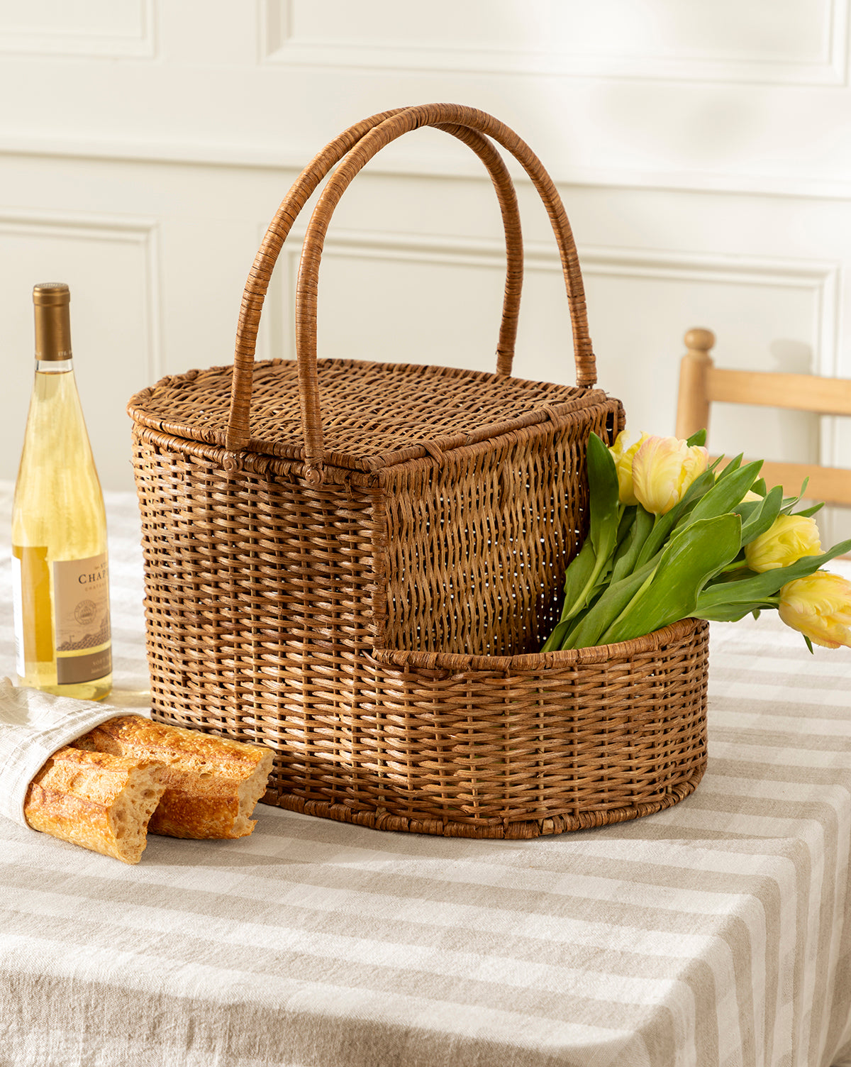 A Rattan Wicker Picnic Basket rests on a striped tablecloth beside a bottle of white wine and a baguette, with a wooden chair and white paneled wall behind, creating an inviting setting for this charming storage piece, by McGee & Co.