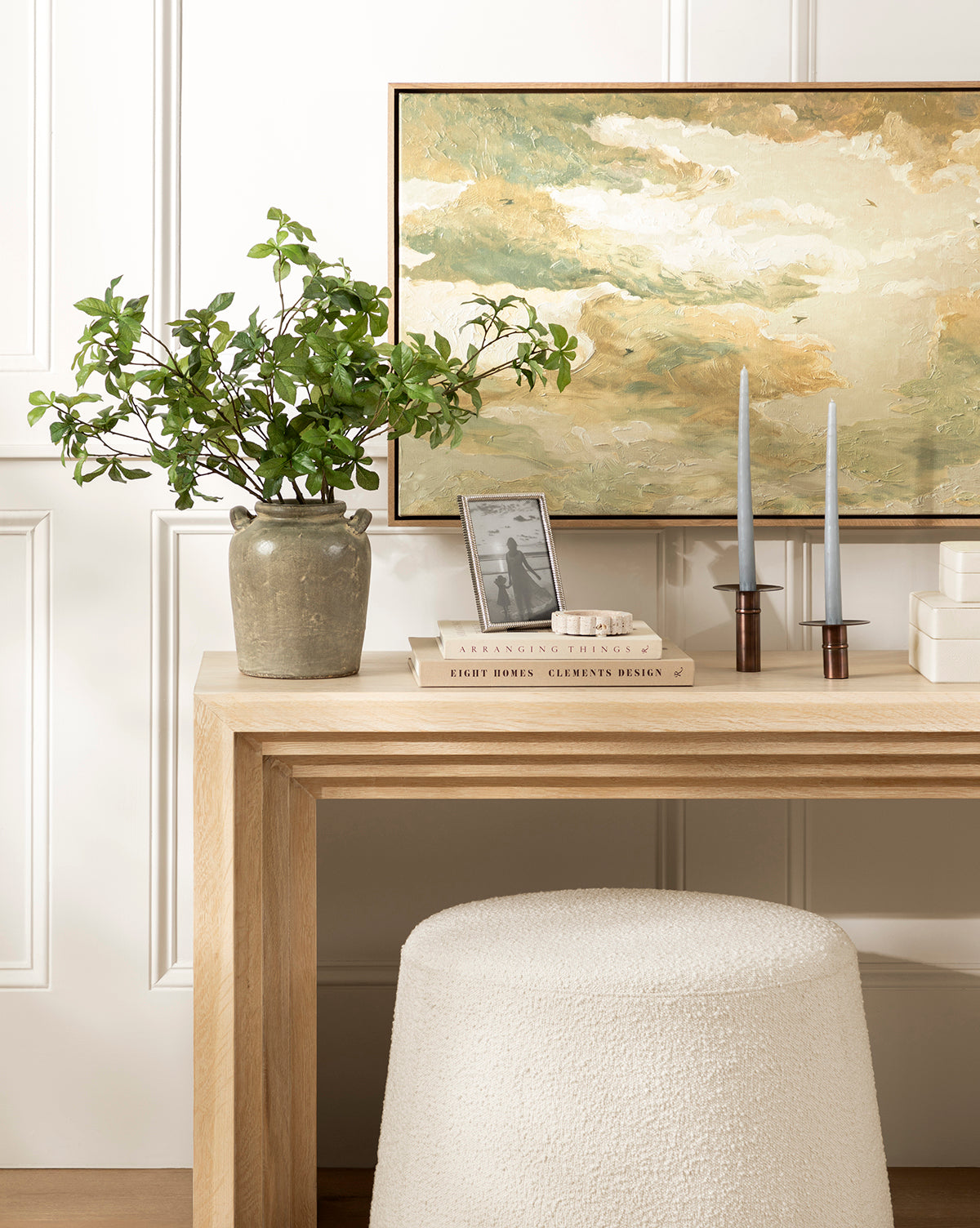 A light wood console table displays a potted leafy plant, Swallows at Dusk wall art in a white oak frame, two blue taper candles in holders, stacked books, and a small photo frame; a textured white stool sits beneath, by McGee & Co.