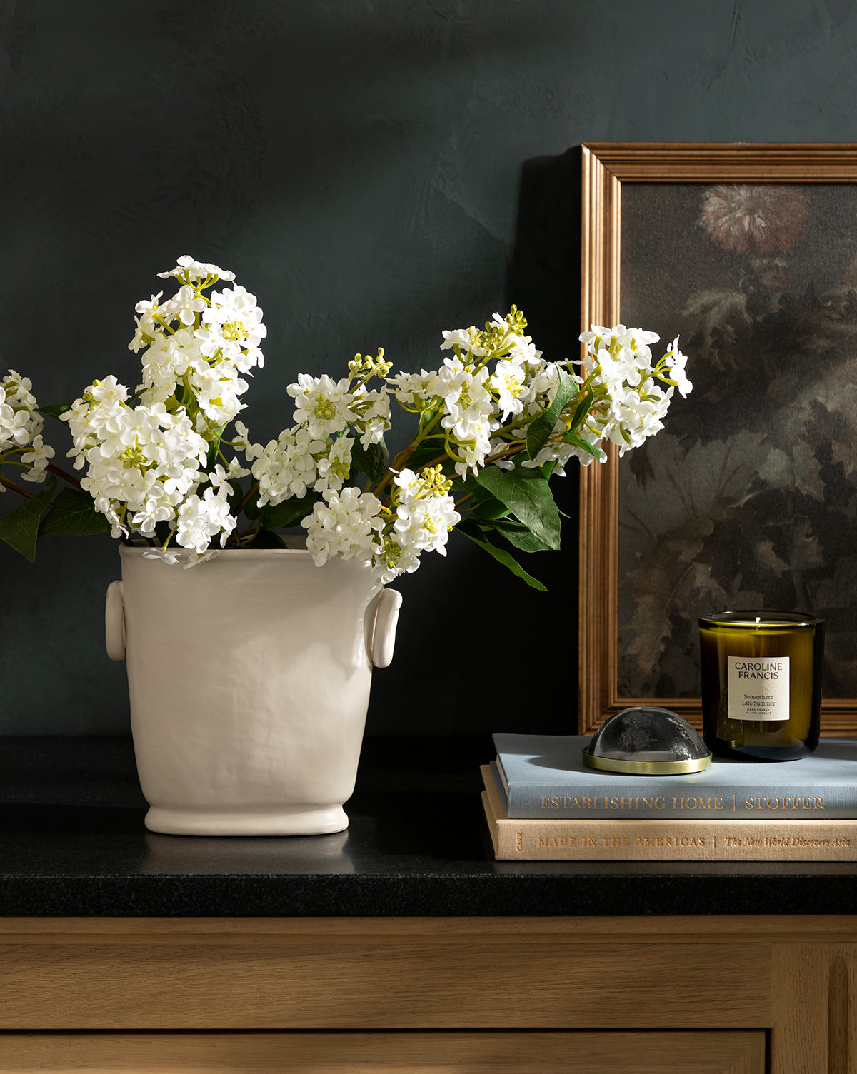 The Powell Ceramic Vase, filled with white flowers, rests on a black countertop beside stacked books, a glass paperweight, a green candle, and a framed photo against a dark wall—an elegant accent for any space.