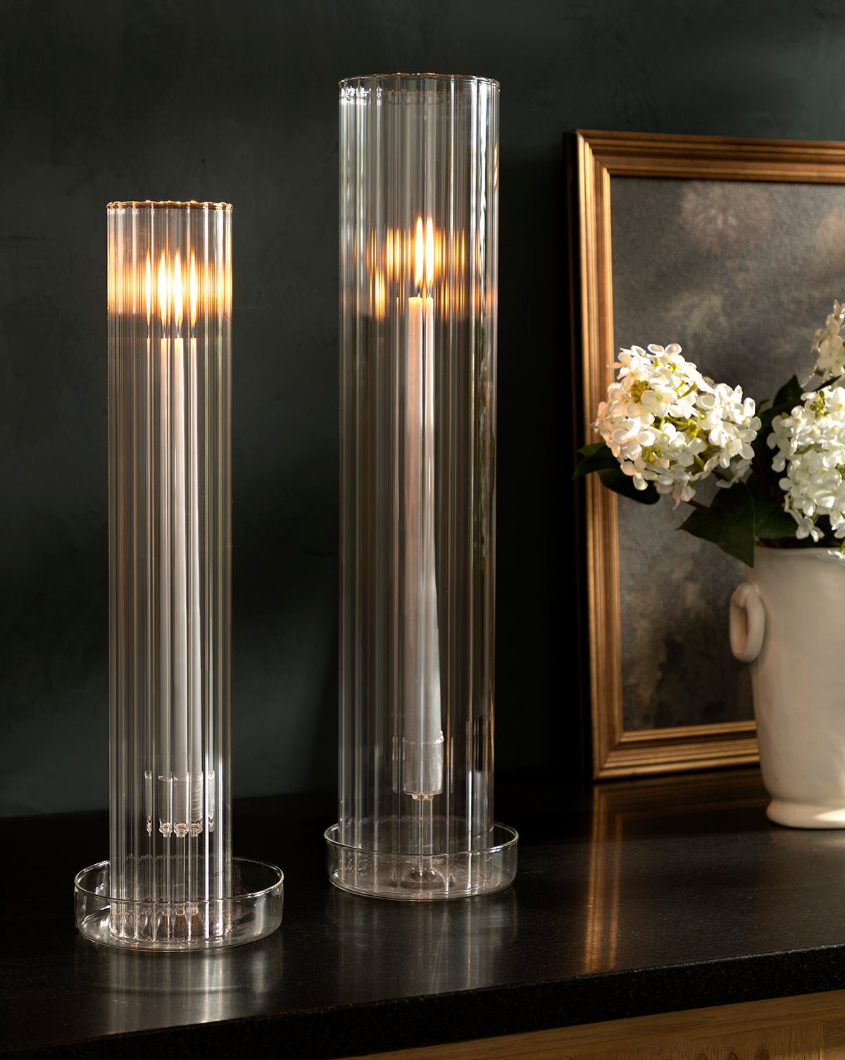 Two lit white candles with Kalina Ribbed Candle Sleeves in elegant glass taper holders sit on a dark surface next to a framed painting and a white vase filled with white flowers.