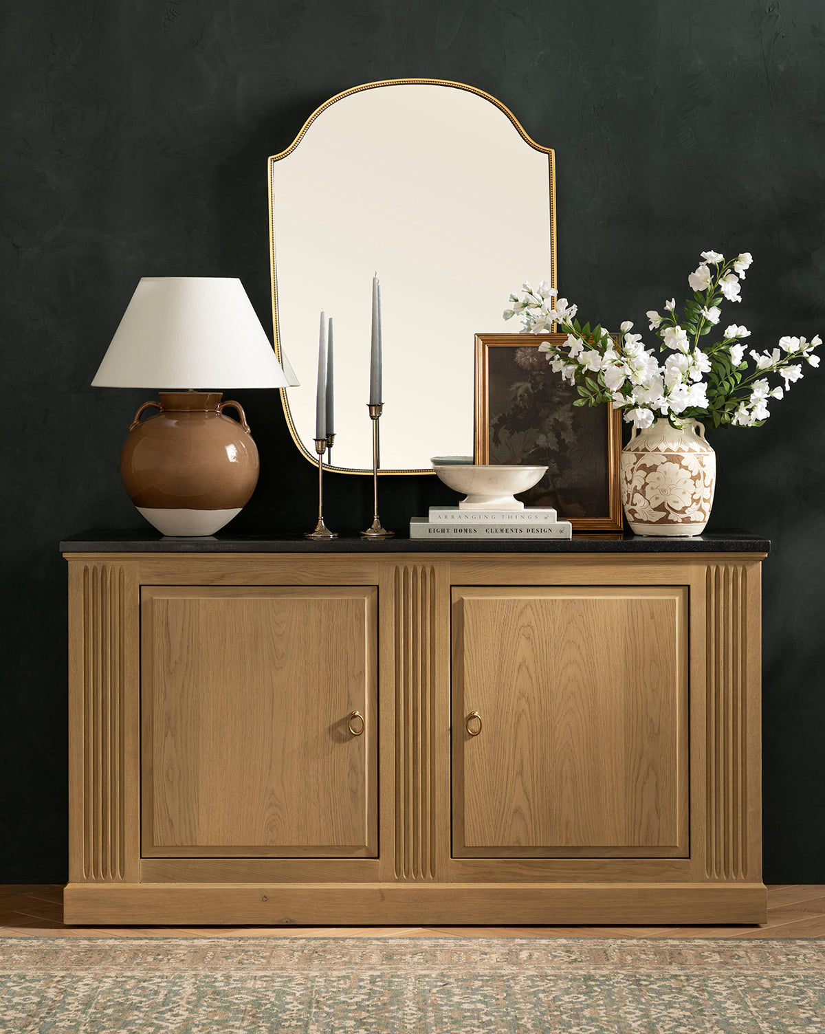 The Ambrose Sideboard by YANG CHENG WOODEN INDUSTRIES INTERNATIONAL (VIETNAM) CO LTD features two doors and is styled with a brown lamp, candles, books, a bowl, framed art, a white flower vase, and a gold-edged mirror above. McGee & Co.