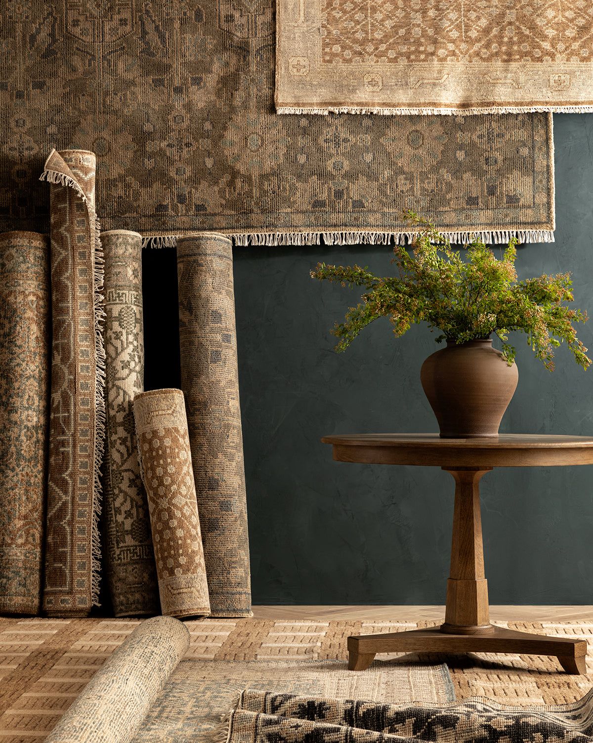Several Endicott Hand-Tufted Wool Rugs in neutral patterns are rolled and hanging against a dark wall, with a wooden table holding a brown vase of green foliage placed in front, atop a textured rug floor by McGee & Co.