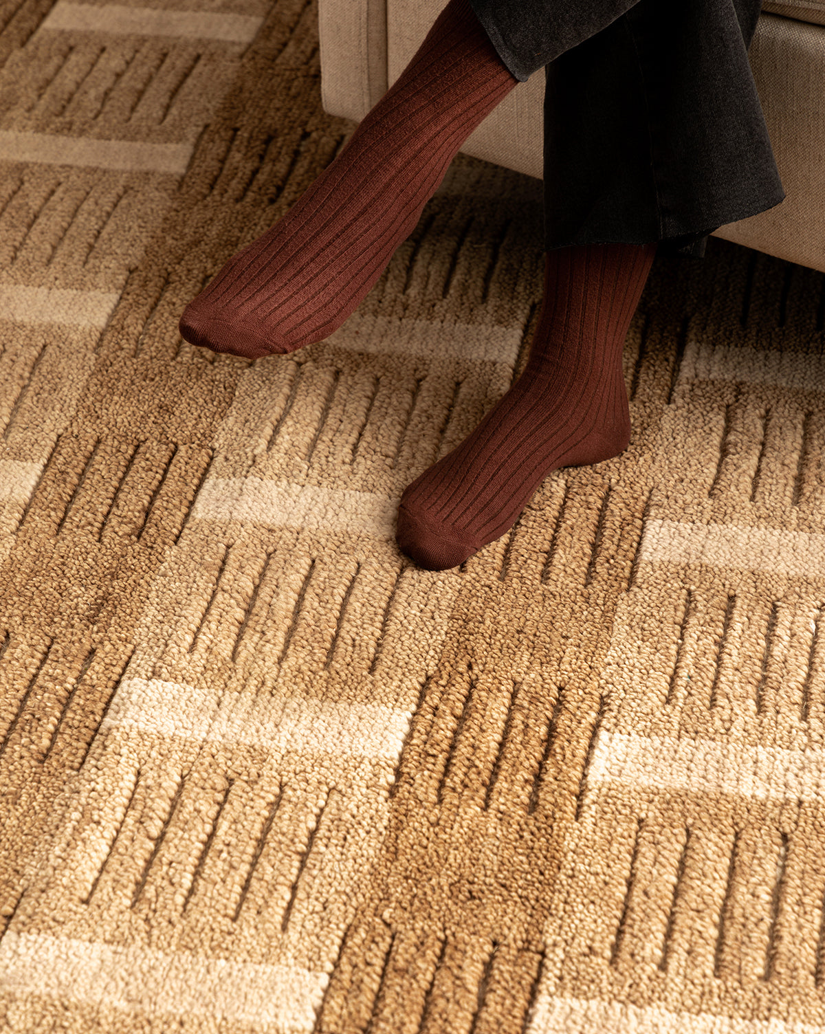 A person wearing dark brown ribbed socks and black pants sits with feet resting on the Brenner Hand-Knotted Wool Rug, which features a beige and tan geometric rectangular pattern, by McGee & Co.