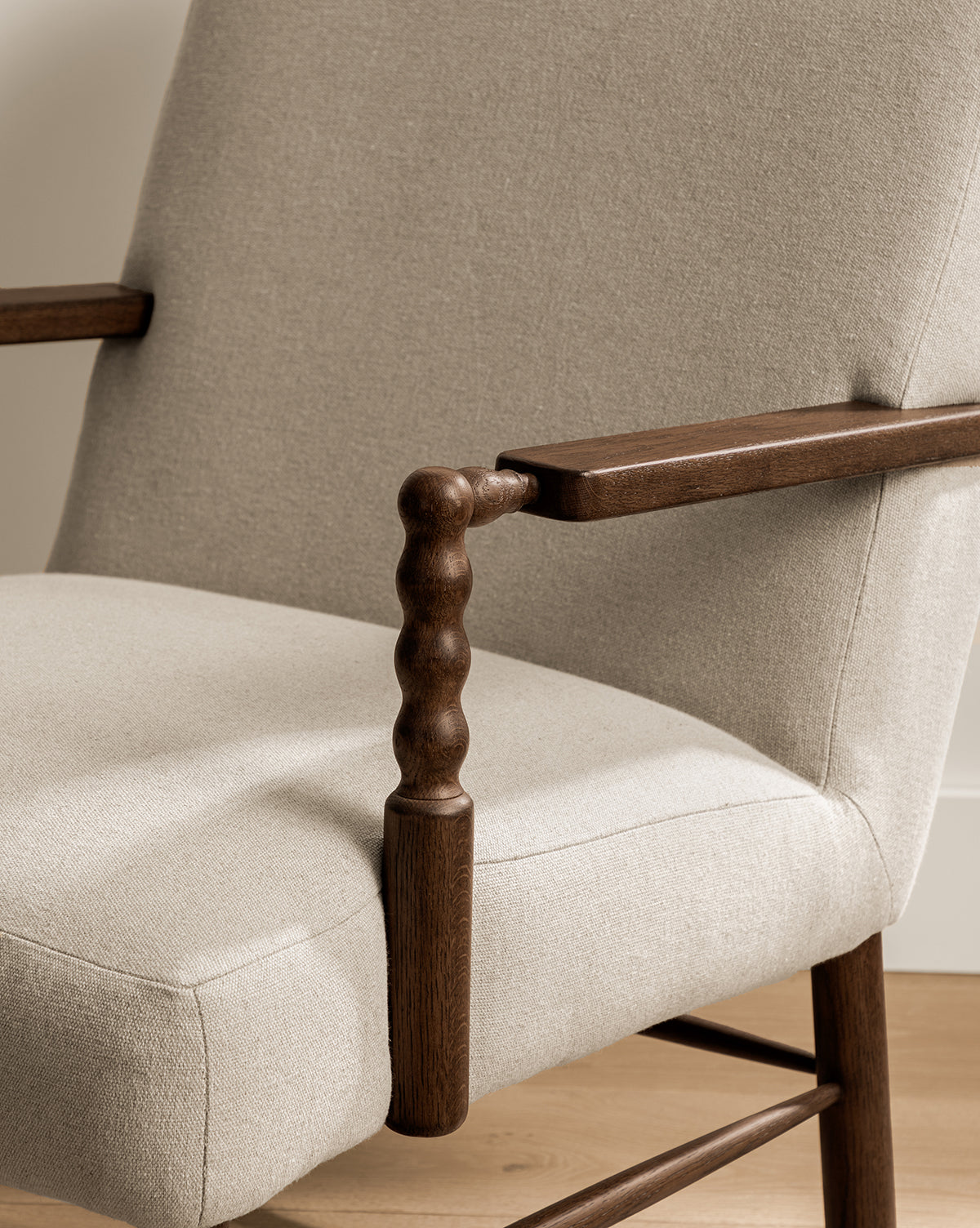 A close-up of the Earnshaw Lounge Chair highlights its neutral-toned upholstery and carved wooden armrests in an English oak finish, elegantly positioned on a light wooden floor, by McGee & Co.