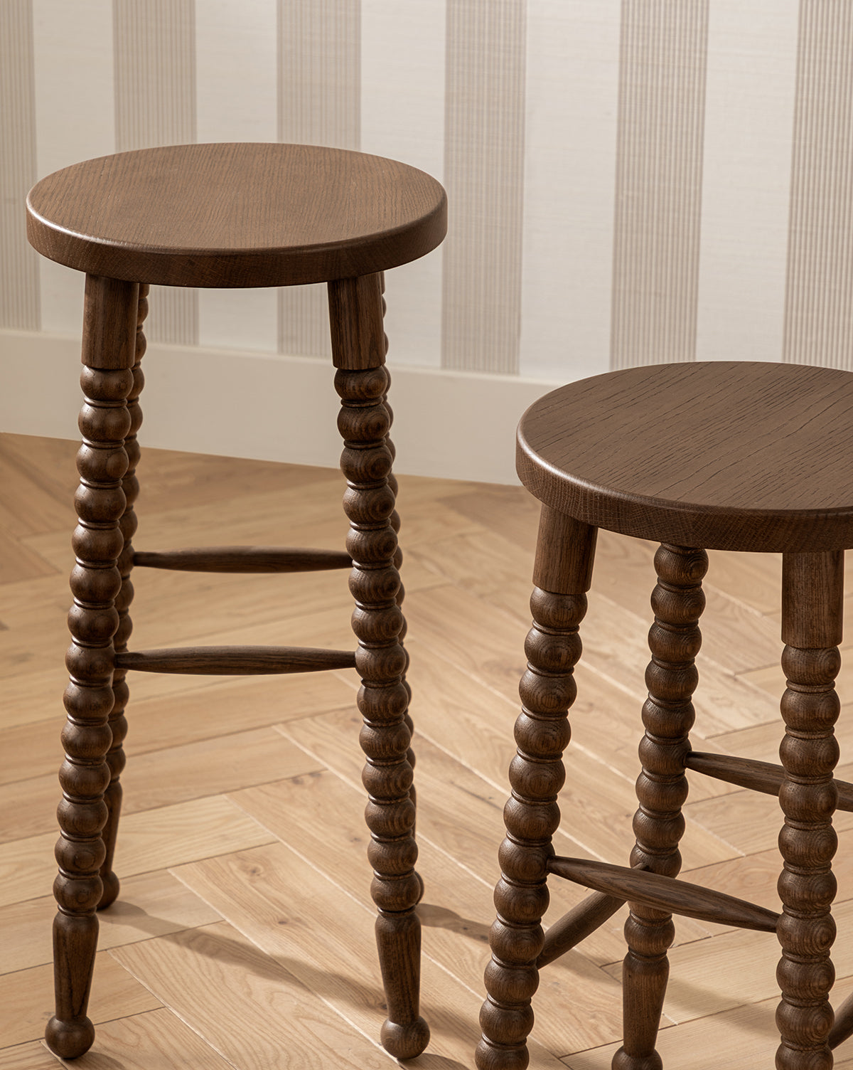 Two Makerspalm Heathcliff Stools with round wooden seats and bobbin-style legs stand on a light herringbone wood floor before a cream wall with striped paneling. McGee & Co.