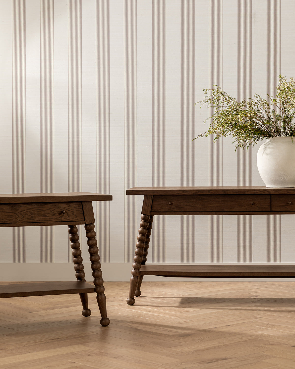 Two Heathcliff Console tables with spindle legs sit on a light herringbone wood floor near a beige and white striped wall. A large white vase with green foliage is placed on the right console, by McGee & Co.