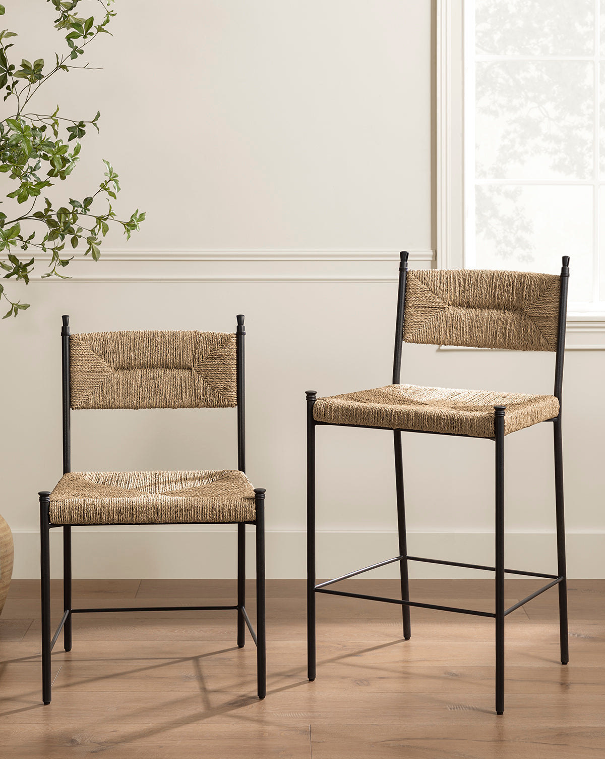 Two Makerspalm Peregrine Stools, featuring black metal frames and woven seagrass seats and backs, sit on a wooden floor near a window and leafy plant in a bright, modern room. One stool is taller for kitchen or dining use. McGee & Co.