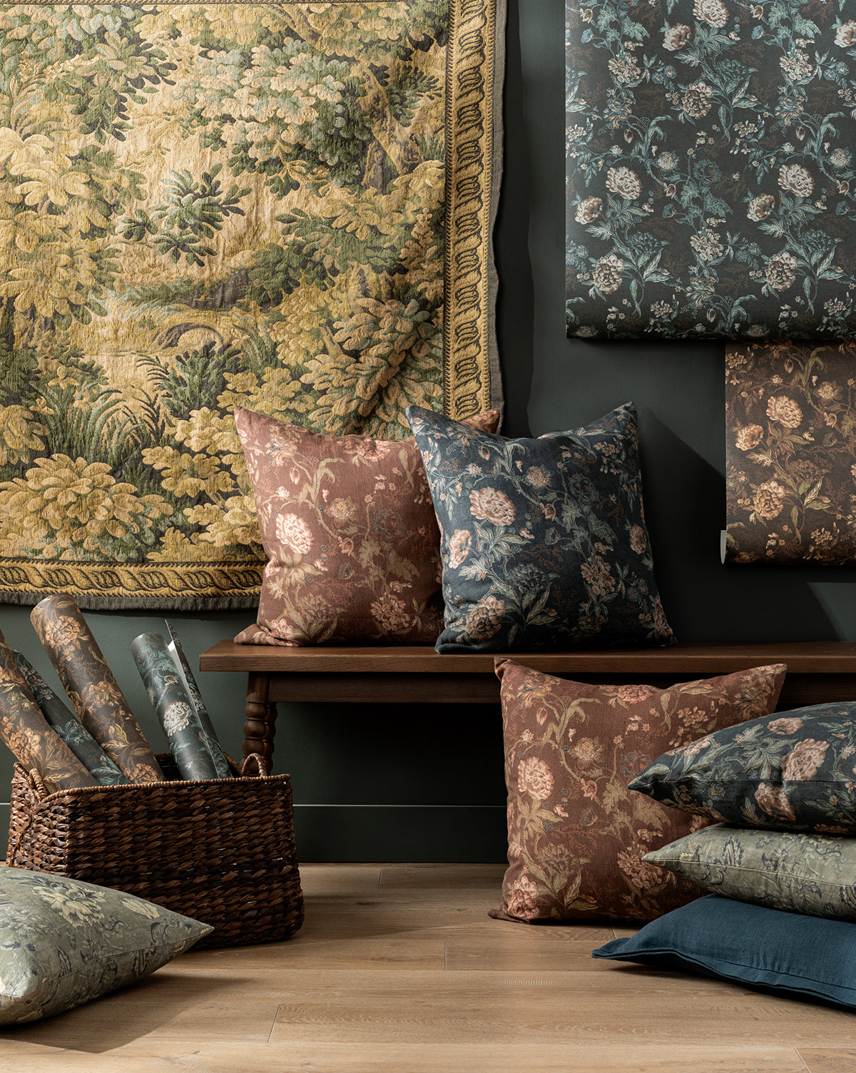 A cozy room with vintage flair: Hestercombe Pillow Covers, patterned pillows, fabric rolls in a basket, a wooden bench, and floral tapestries—styled in earthy green, blue, and brown tones by McGee & Co.