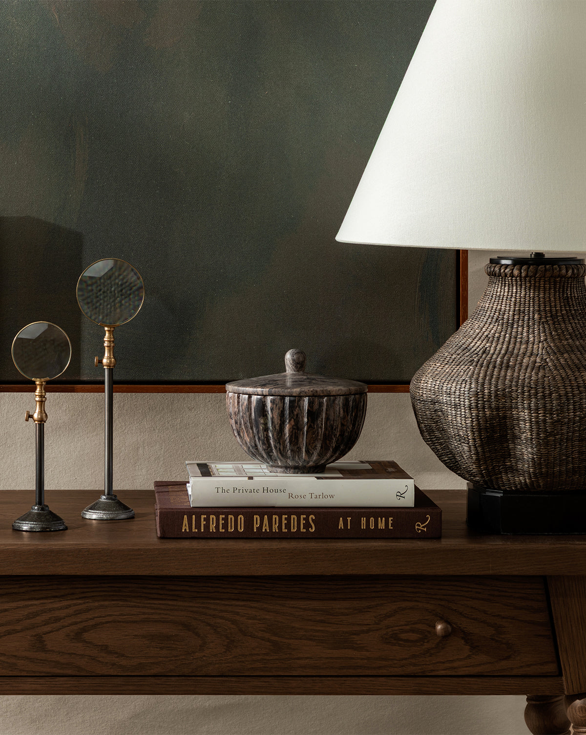 A wooden console table displays two stacked books, magnifying glasses on stands, a lidded bowl, and the Tahiti Table Lamp with a linen shade, all set before greenish wall art by McGee & Co.