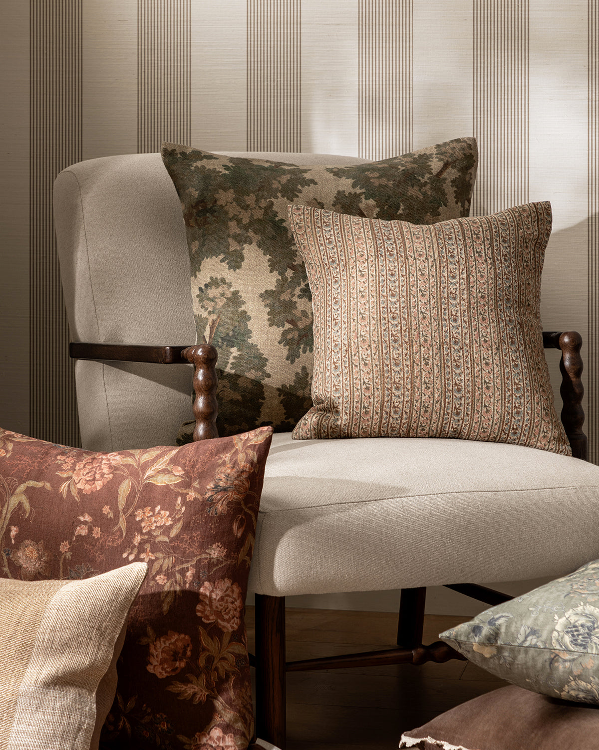 A beige upholstered chair holds two pillows—one adorned with the Wakefield Pillow Cover, featuring a leafy camouflage design, and another with stripes. In front, more floral and patterned pillows in neutral tones are artfully arranged by McGee & Co.