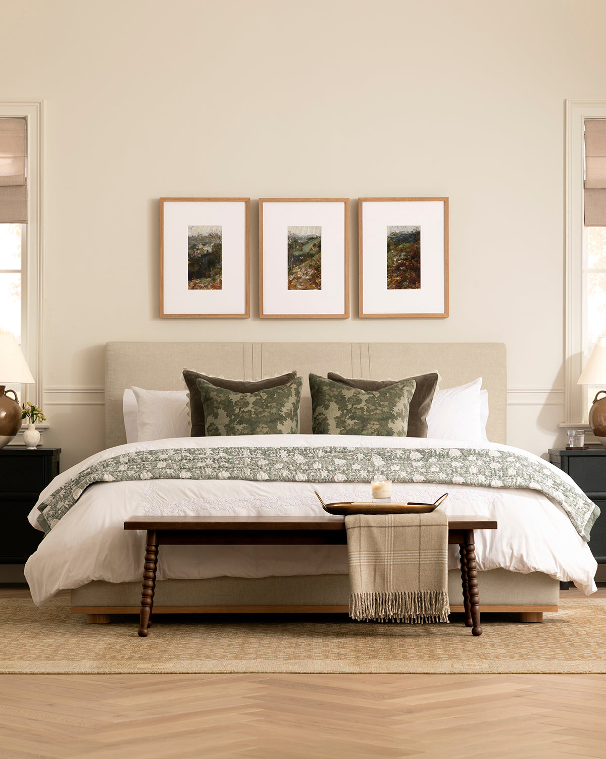 A neatly made bed with white and green bedding, three green pillows, and a Heathcliff Bench at the foot holding a tray and blanket; three framed artworks hang above, while bedside tables with lamps are on each side, by McGee & Co.