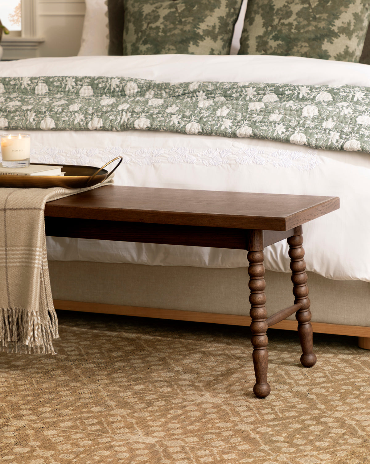 A Heathcliff Bench sits at the foot of a bed with green and white bedding; a tray holding a candle and book, along with a beige throw blanket, are arranged on the bench, by McGee & Co.
