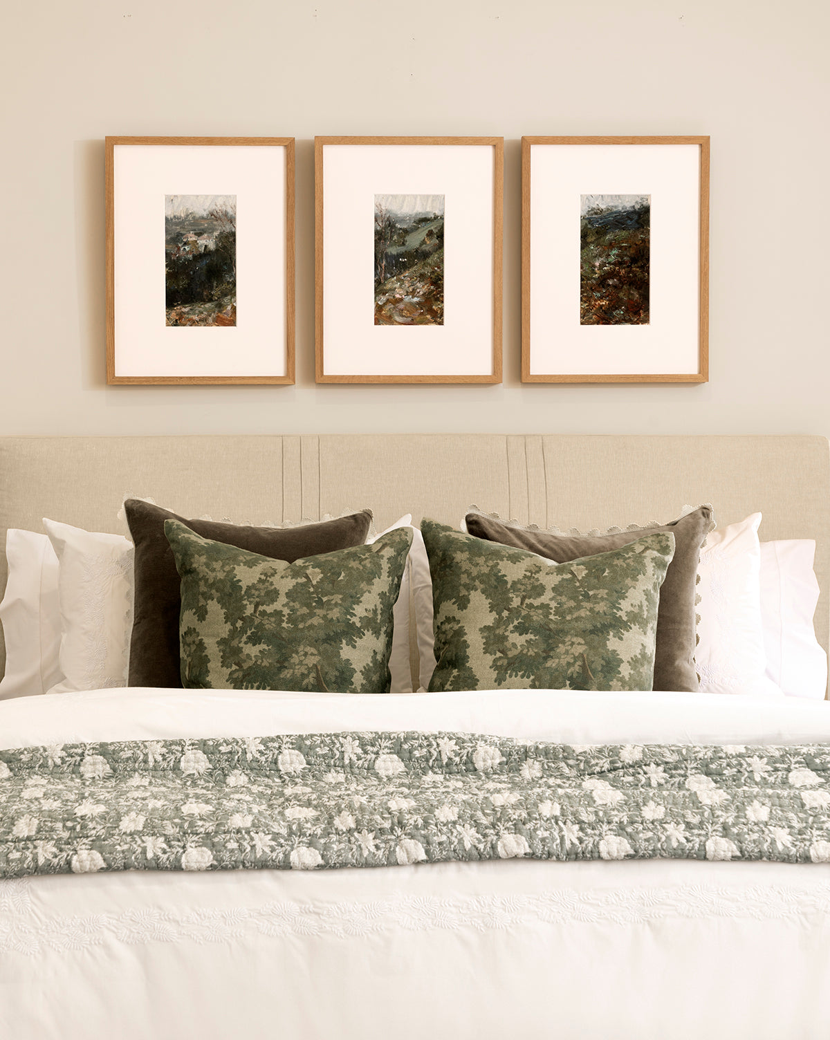A neatly made bed with white linens and green pillows sits beneath A River Runs Through, a set of three framed abstract landscapes, adding striking wall art above the beige headboard on the light wall, by McGee & Co.