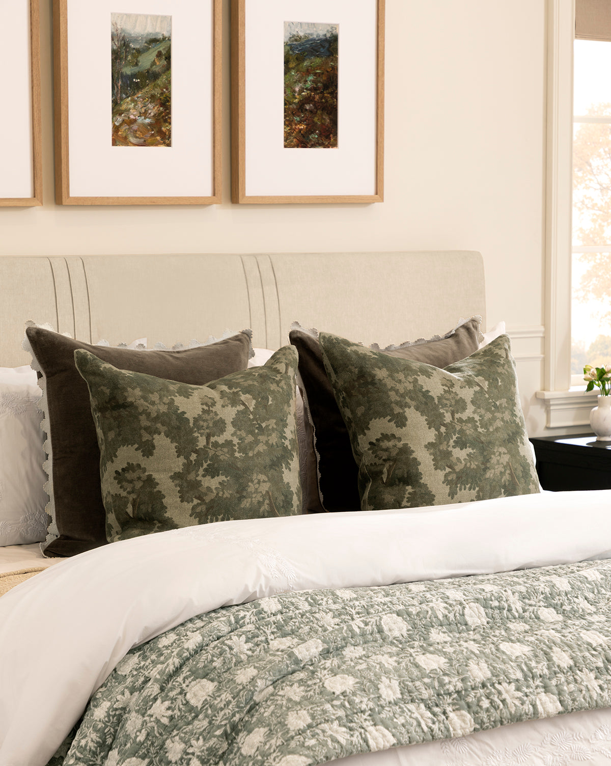 A neatly made bed features green and white floral bedding, two Lucetta Pillow Covers with a leafy pattern in earthy tones, two solid dark green pillows, and three framed landscape artworks above on a light-colored wall, by McGee & Co.