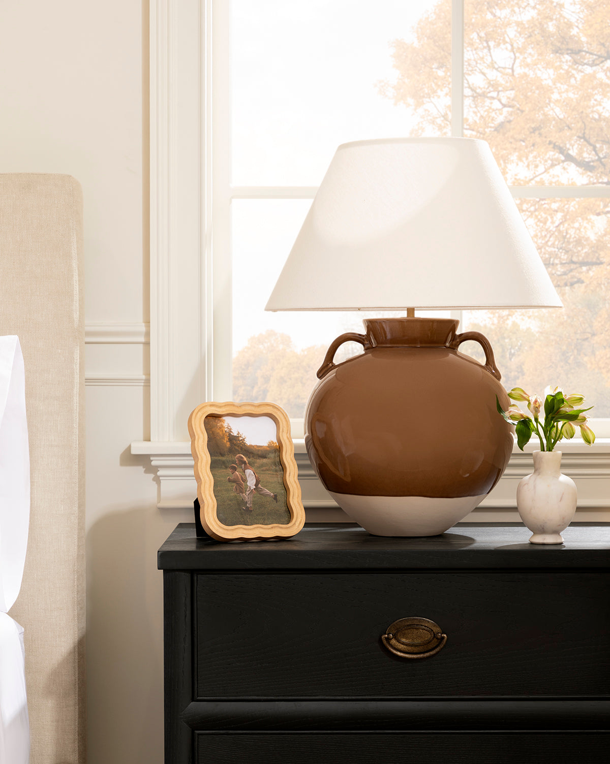 A natural rubberwood bedside table with a dark drawer displays a large brown lamp with a white shade, yellow flowers in a vase, and a photo in the Scalloped Wood Frame. The room includes a bed and window showing autumn trees outside, by McGee & Co.