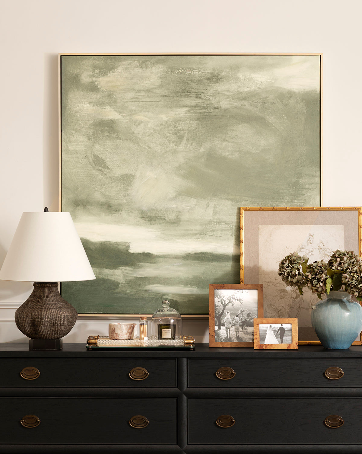 A dresser with brass handles holds a Tahiti Table Lamp, framed photos, a tray with glass items, dried flowers in a blue vase, and several framed artworks. A large green abstract painting hangs on the wall above, by McGee & Co.