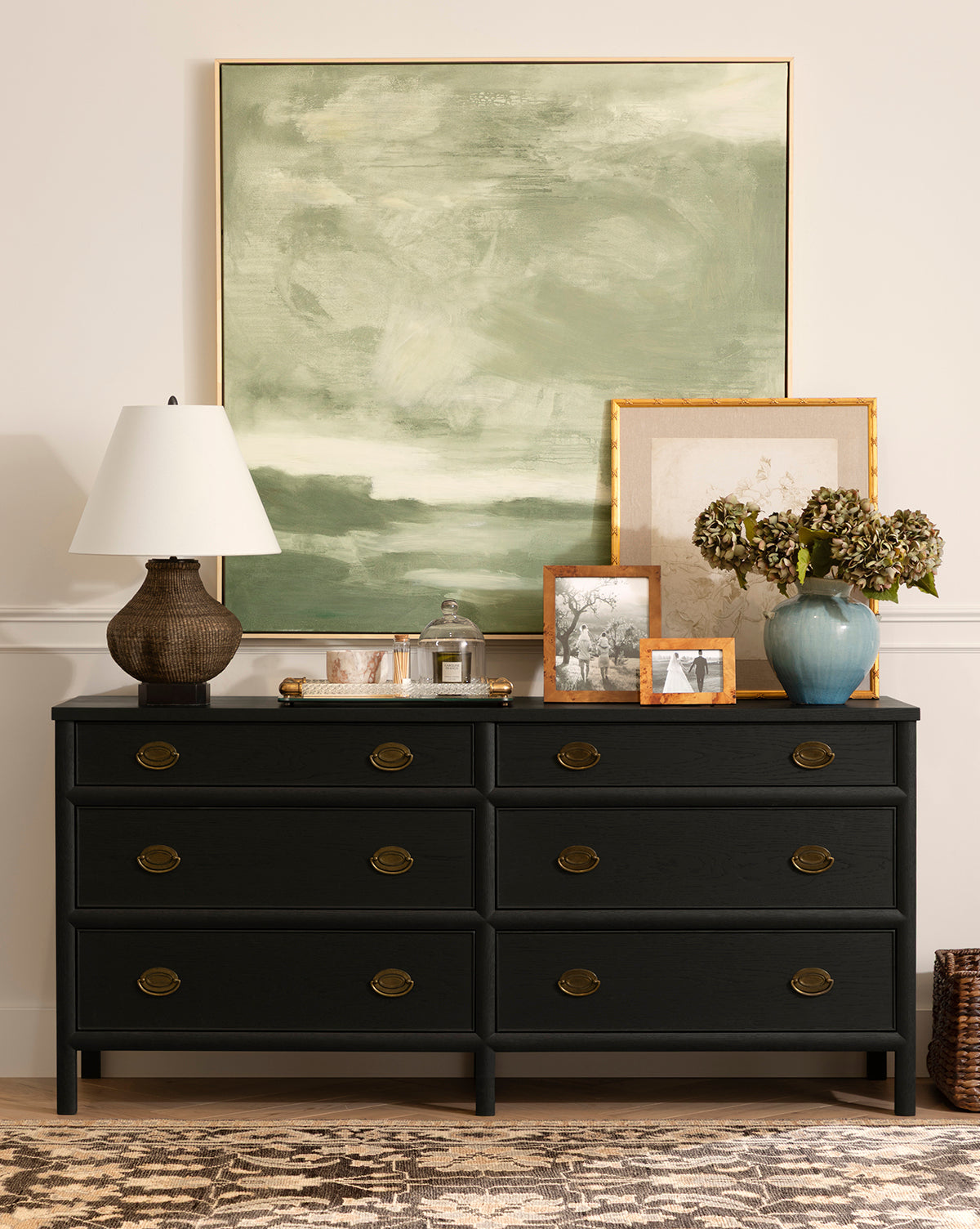 The Edward Dresser in Black Ash with brass knobs provides chic bedroom storage, adorned with a textured lamp, tray, glass jar, framed photos, vase of dried flowers, and two large overlapping art pieces above a patterned rug, by McGee & Co.