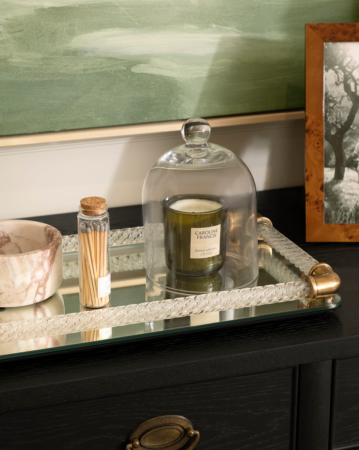 A glass cloche covers a green candle on a mirrored tray beside the Verona Marble Bowl, which has brown veining and a matte finish. A glass jar of matches, a framed black-and-white photo, and a green painting complete the elegant scene by McGee & Co.