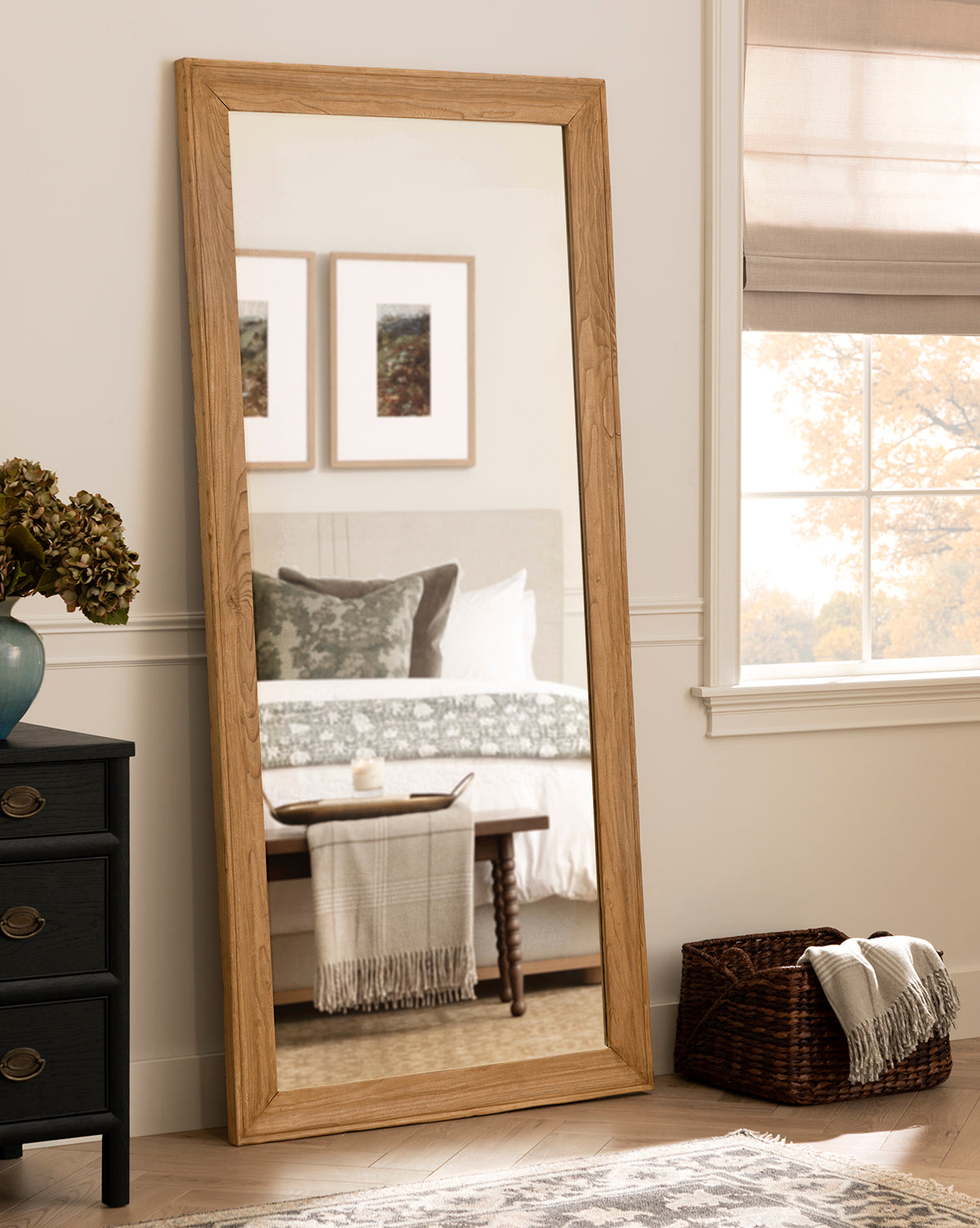The Pippen Floor Mirror, featuring a honey elm finish, leans against the wall and reflects a bed with green and white bedding, a bench with a striped throw, two framed pictures, plus a nearby black dresser and window with beige blinds by McGee & Co.