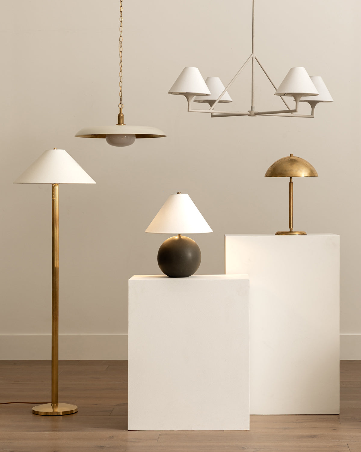 The Townes Floor Lamp in gold reeded brass, a black table lamp, and a gold desk lamp stand on white pedestals; above are a white linen-shade chandelier and a gold pendant, all set against a neutral backdrop, by McGee & Co.