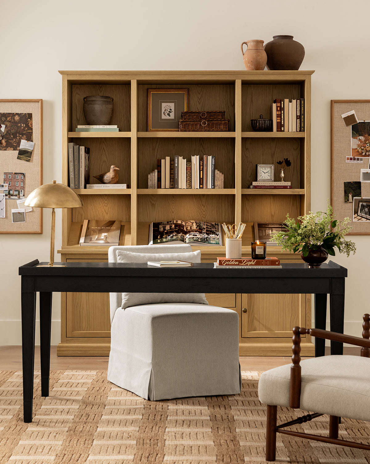 A cozy home office features a wooden bookcase, black desk with lamp, books, candle, vase of flowers, cushioned chair, cork boards on a cream wall, and the Brenner Hand-Knotted Wool Rug grounding the space, by McGee & Co.